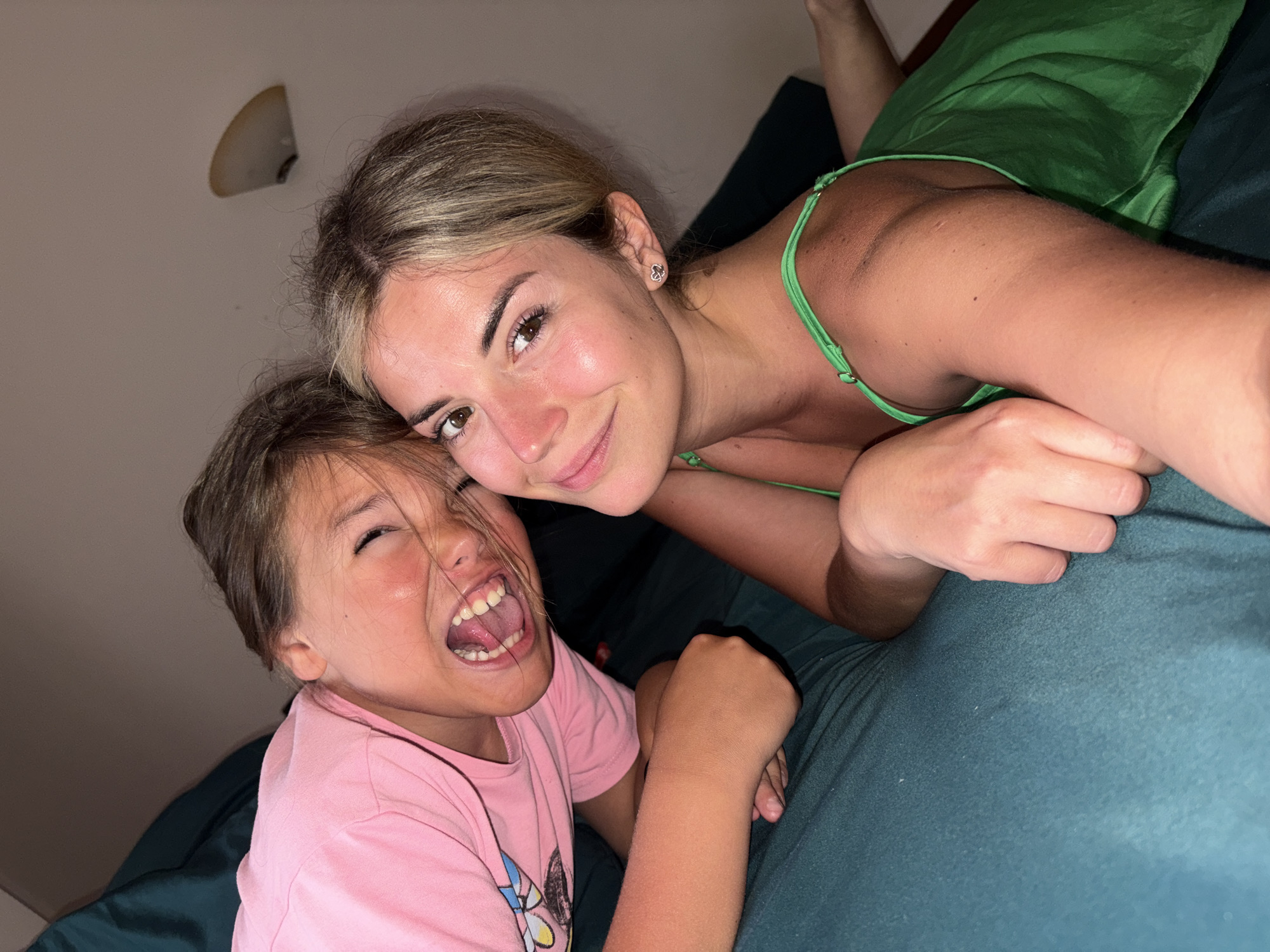 Two females, one younger and one older, are lying on a teal-colored surface that appears to be a bed. The younger female is on the left, wearing a pink t-shirt, and is laughing with her mouth wide open. Her head is tilted back, and her eyes are closed. The older female is on the right, wearing a green spaghetti strap dress, and is looking at the camera with a slight smile. Her blonde hair is pulled back, and she is wearing small heart-shaped earrings. Her left arm is resting across the younger female's back. The background is dimly lit and shows a wall with a wall-mounted light fixture. The scene suggests an indoor environment, possibly a bedroom. The image was taken indoors in Budva, Montenegro.