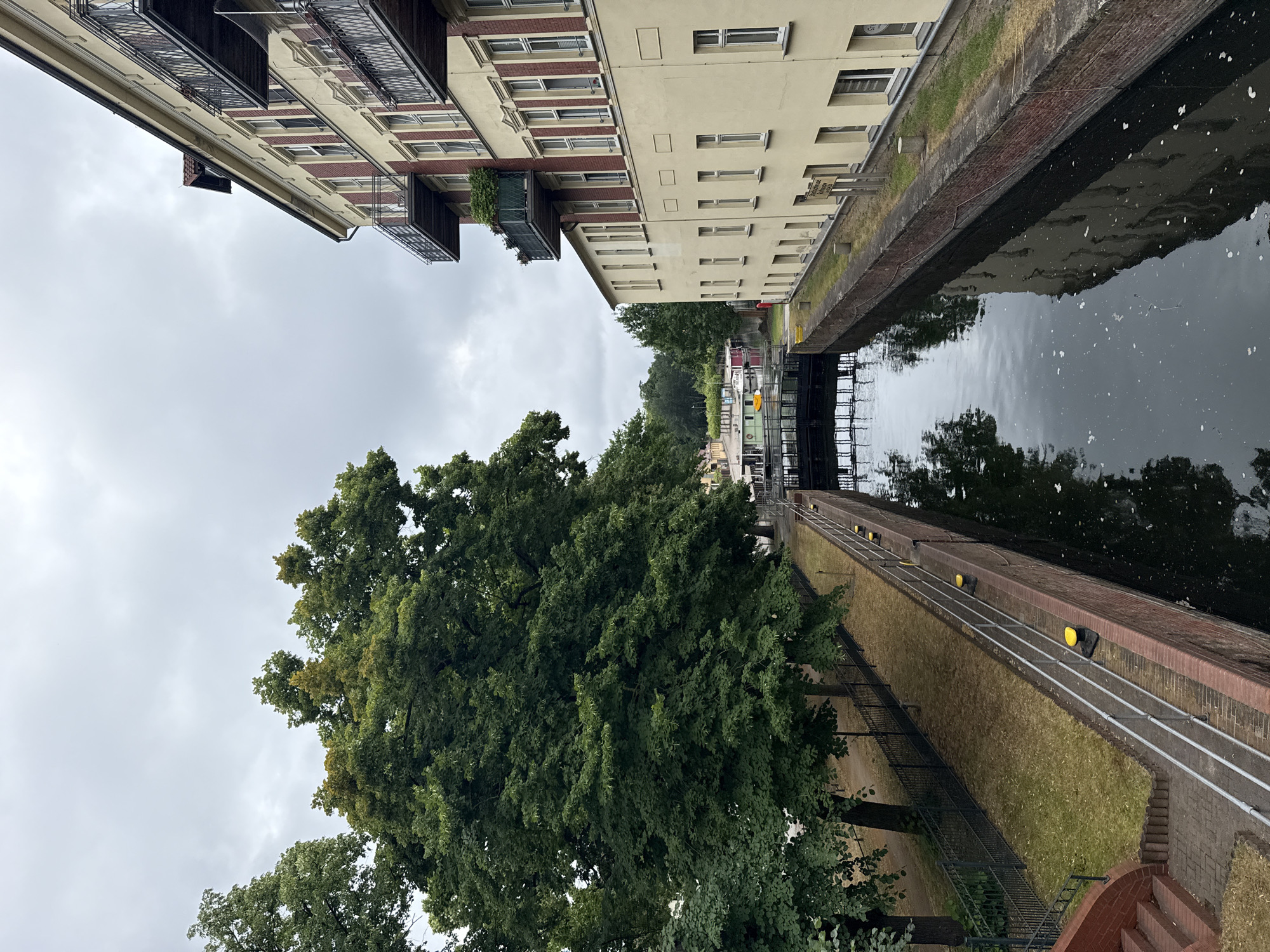 The image shows a canal in the city of Rathenow, Germany, with a large building on the right and trees on the left. The canal has brick walls with metal railings and yellow bollards. A lock gate is visible in the distance. The building on the right is a multi-story structure with a yellow facade, red brick accents, and balconies. The trees on the left are lush and green, with dense foliage. The sky is overcast and cloudy. The water in the canal reflects the sky and the building. In the distance, there are some smaller boats moored. The scene depicts a serene urban waterway with a mix of architectural and natural elements.