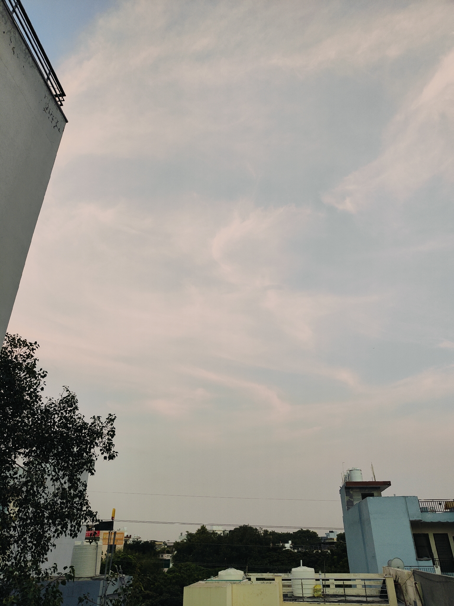 A low-angle, wide shot captures a sky filled with wispy, light pink and gray clouds. To the left, the corner of a tall, light-colored building juts into the frame, its top edge marked by a dark railing. A dark green tree with dense foliage extends from the lower left corner, obscuring part of the building. In the middle ground, a line of trees forms a dark, green horizon. On the right side of the image, a light blue building with a dark railing on its upper story rises above the trees. A white water tank sits atop this building, and a red-capped water tank is visible beside it. Several other water tanks and parts of buildings are visible in the middle ground and background. The scene appears to be an urban residential area in Gurgaon, India, possibly in the late afternoon or early evening given the cloud colors and lighting.