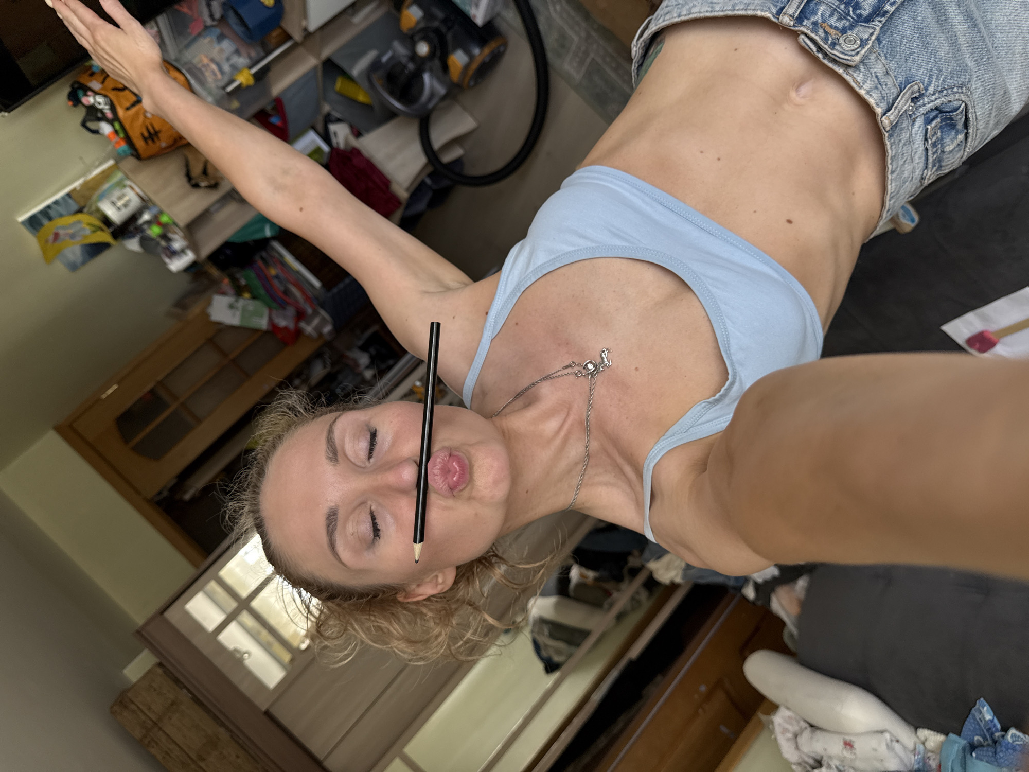 A selfie captures a blonde woman indoors. She is angled, holding a pencil horizontally in her mouth and eyes closed. She is wearing a light blue tank top and a denim jacket. A silver chain necklace is visible. The background includes wooden furniture, a vacuum cleaner, and various scattered items. The location is an interior space, likely in an apartment or house. There is no evidence indicating that the location is in the city of Chaikovsky, Russia.