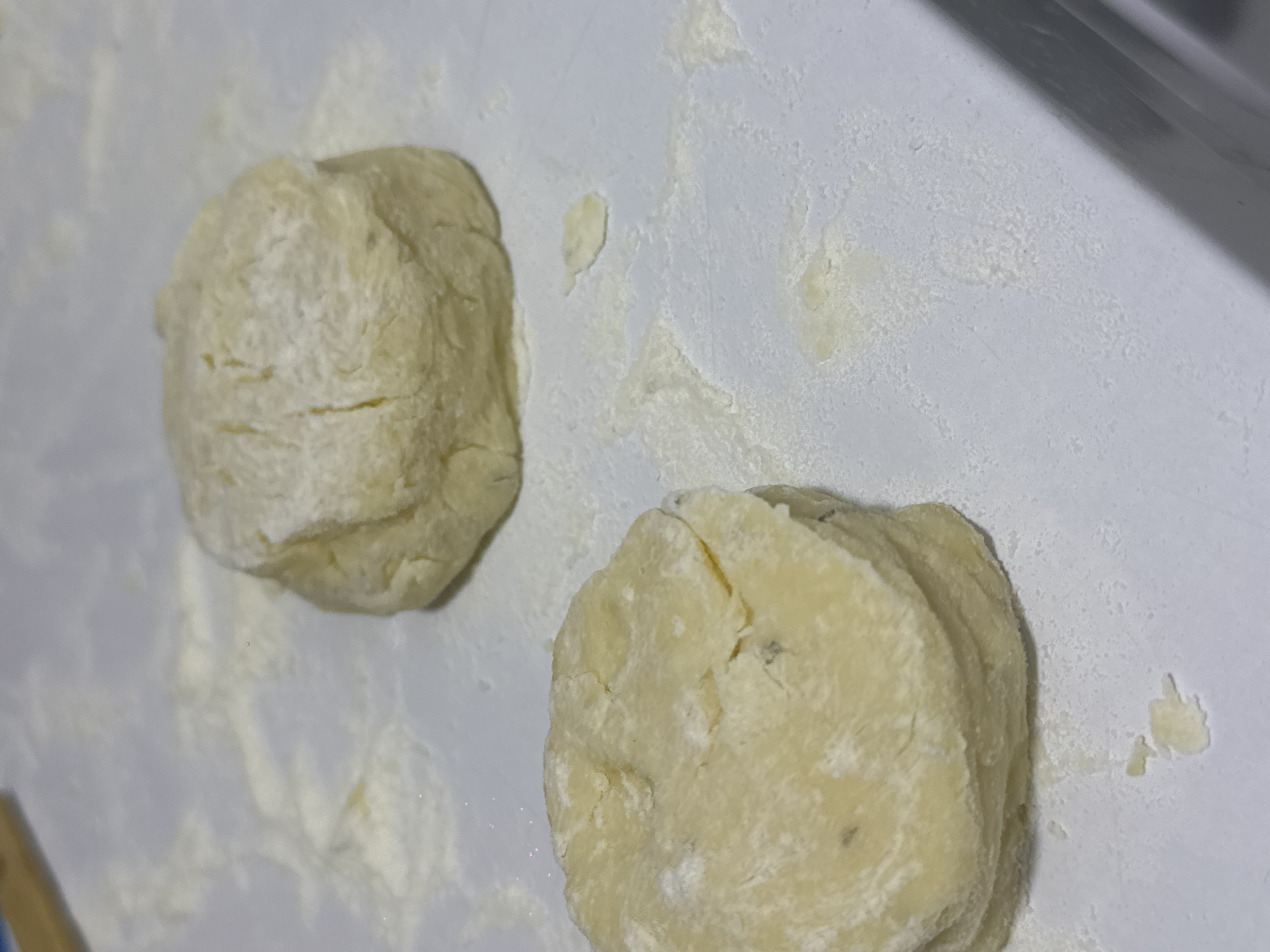 Two mounds of uncooked biscuit dough are on a white surface dusted with flour. The dough is pale yellow and appears to have been cut into rounds or folded. One mound, in the foreground on the left, is stacked, showing several layers. The other mound, in the background on the right, is a single, more irregular shape. Flour is scattered around the dough and on the surface, which is a light color and shows some faint smudges and textures.