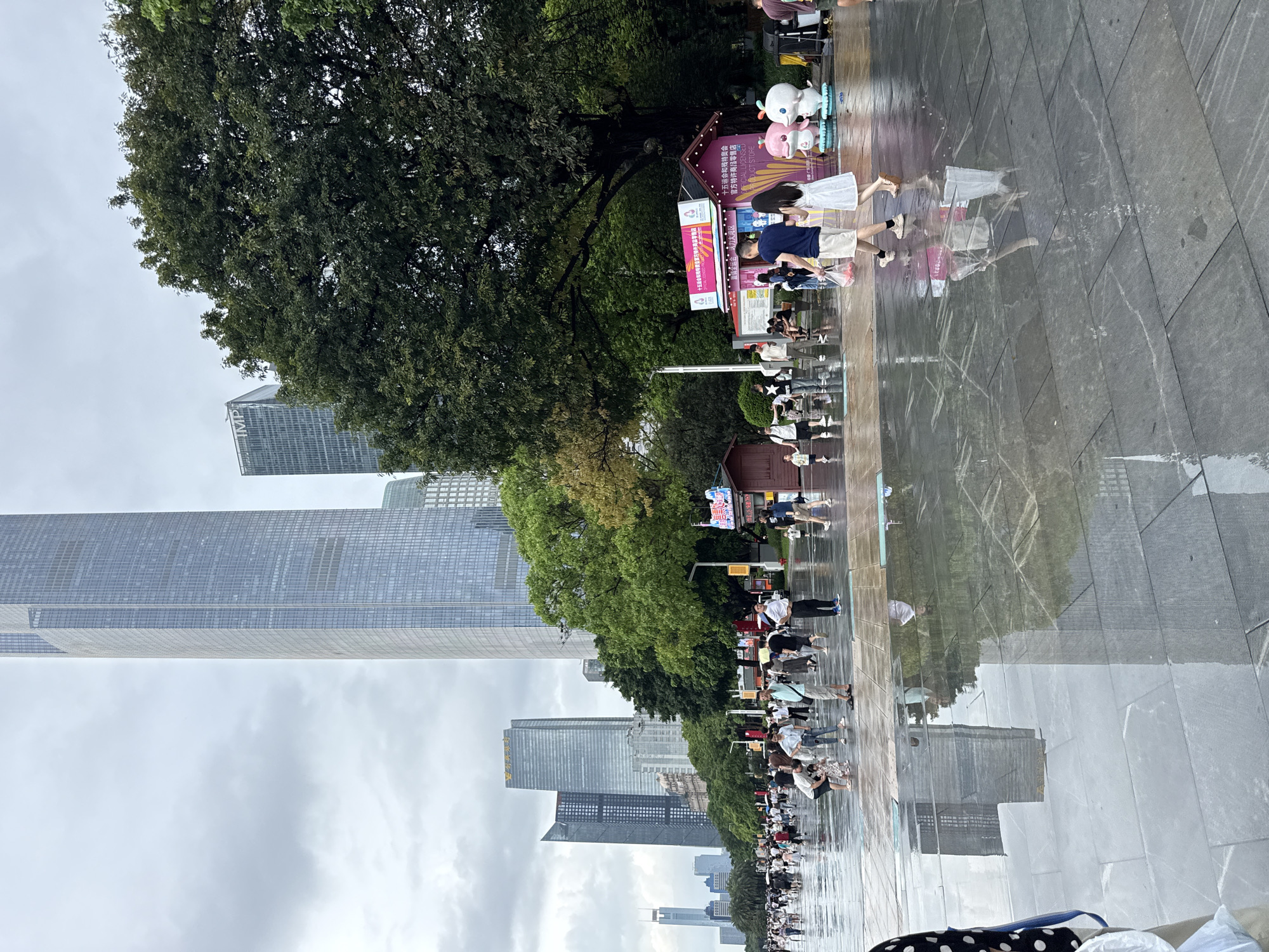 The image depicts a city scene in Tianhe District, China, on a cloudy day. The foreground is a wet, paved plaza with reflections of the sky and surrounding buildings. Several people are walking on the plaza, some singly and some in small groups. They are dressed in casual summer clothing, and some are carrying shopping bags.
In the background, there are several tall, modern skyscrapers, including one with a distinctive pointed top and another with "IMP" visible on its facade. Lush green trees are scattered throughout the cityscape, providing a contrast to the urban architecture.
To the right, a small, pink, booth-like structure with signage and cartoon characters is visible, possibly a vendor or display. The overall impression is of a bustling urban area after rain, with a mix of modern and natural elements.