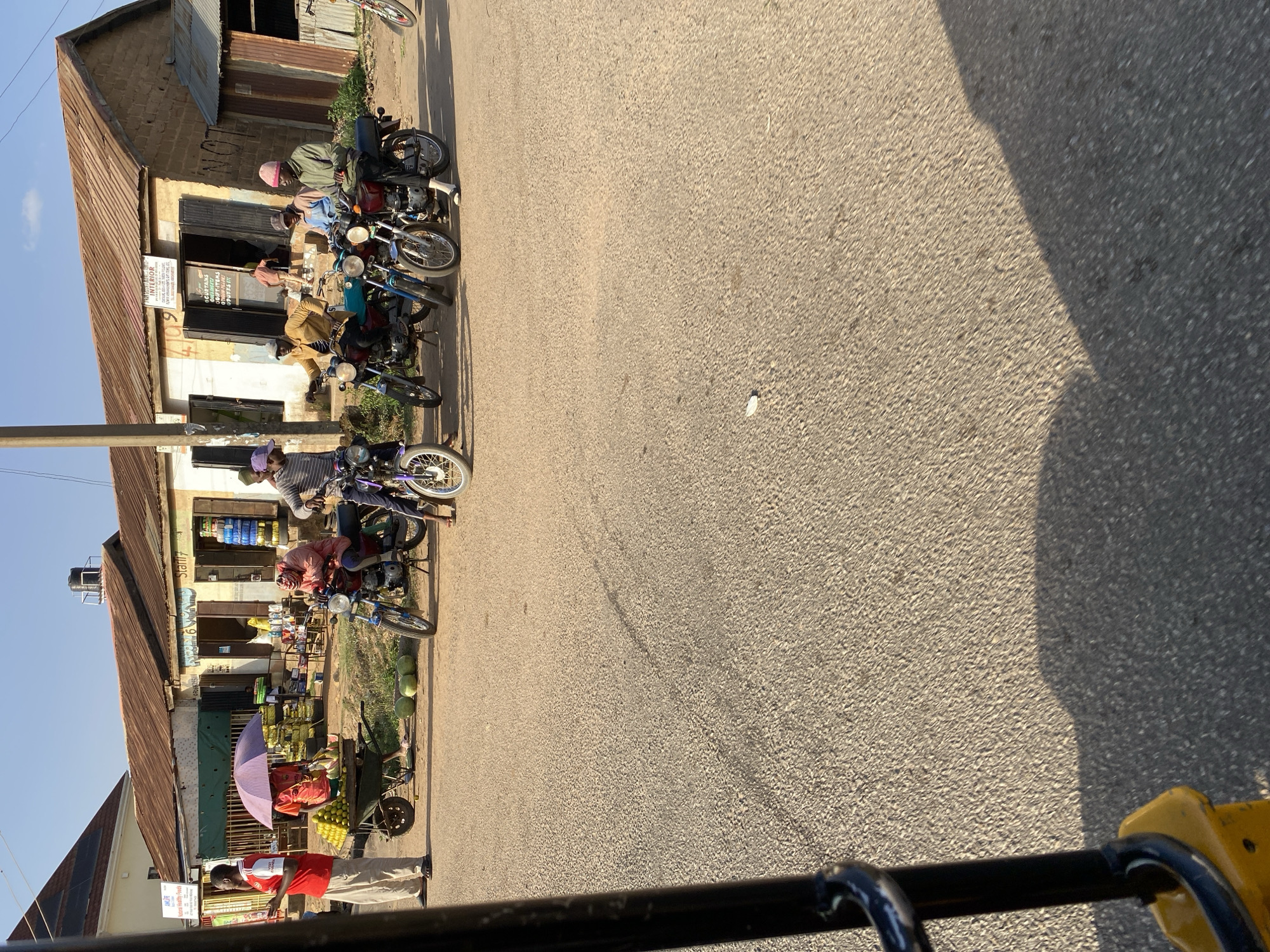 The image shows a street scene in Bukuru, Nigeria. Several motorcycles are parked along the edge of the road in front of a row of small shops. People are seated on some of the motorcycles, while others stand or sit near the shops. A vendor stands in front of a building wearing a red shirt and khaki pants. The buildings are weathered, with corrugated metal roofs. A market or roadside display with an umbrella is partially visible. The road is paved and takes up a large portion of the image. The view is likely from inside a vehicle.