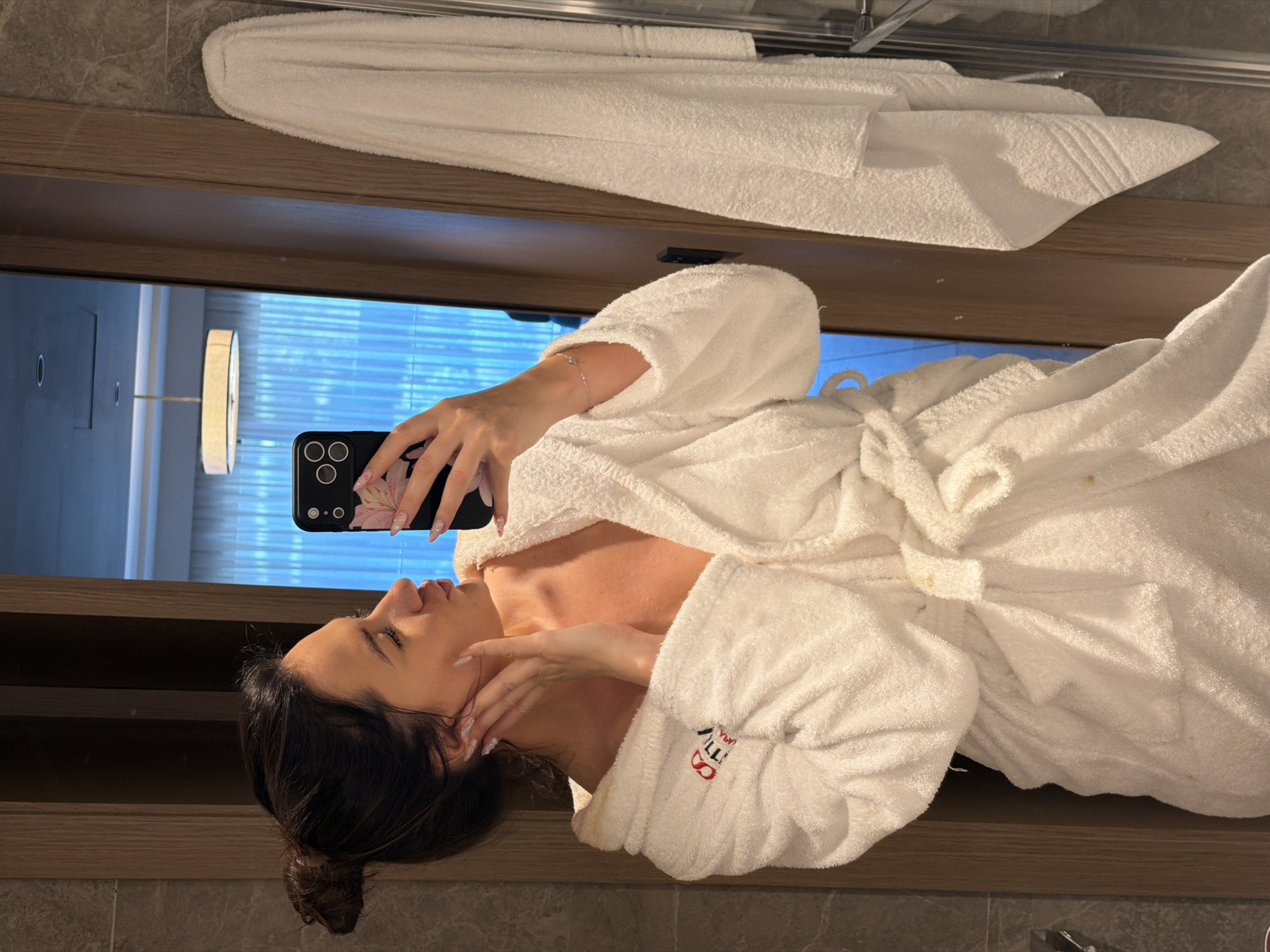 A woman in a white robe is taking a selfie in a bathroom mirror. The robe appears to have a logo, and a white towel is folded above the mirror. Behind her, through the mirror's reflection, is a window with blinds. There is also a light fixture visible in the reflection. The location appears to be a hotel bathroom. It is not possible to confirm the city of Küçükçekmece, Turkey, based on the visual information alone.