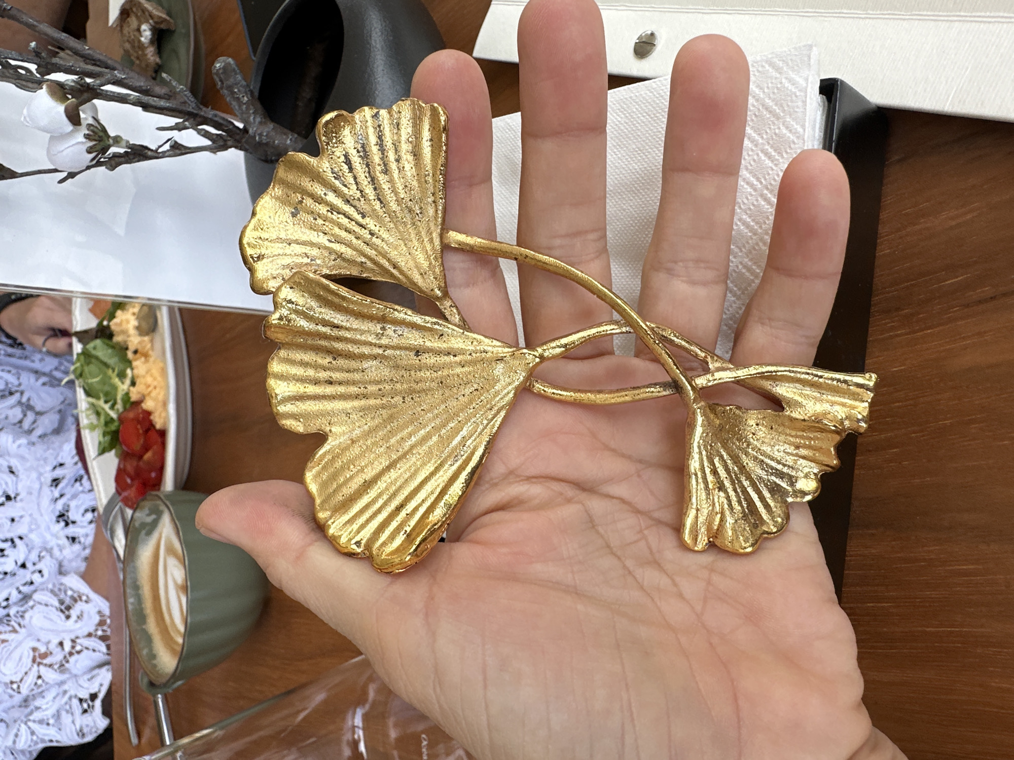 The image captures a person's open hand, palm facing upwards, holding a striking golden decorative item shaped like several ginkgo leaves with intertwining stems. The leaves are textured with radiating veins, giving them a realistic yet stylized appearance. The person's fingers are slightly curled around the stems of the ornament, which features two larger ginkgo leaves and a smaller one.
The setting appears to be a cafe or restaurant, indicated by the wooden tabletop, tableware, and food items. To the left, a plate holds a meal featuring greens, possibly tomatoes or berries, and a yellowish spread or dip. Next to the plate is a green ribbed cup containing a coffee with latte art. A glass of water is also visible on the table.
In the background, a small black vase or container holds a branch adorned with artificial white flowers or buds. Part of a white surface, possibly a napkin or menu, with metallic fasteners, is also seen behind the hand. A portion of another person, wearing white lace clothing, is partially visible to the left of the food plate, suggesting a companion at the table. The lighting suggests it is daytime. No visible text is present in the image. The scene conveys a casual dining moment in ตำบลเกาะพะงัน, Thailand, where a decorative piece is being observed or presented.