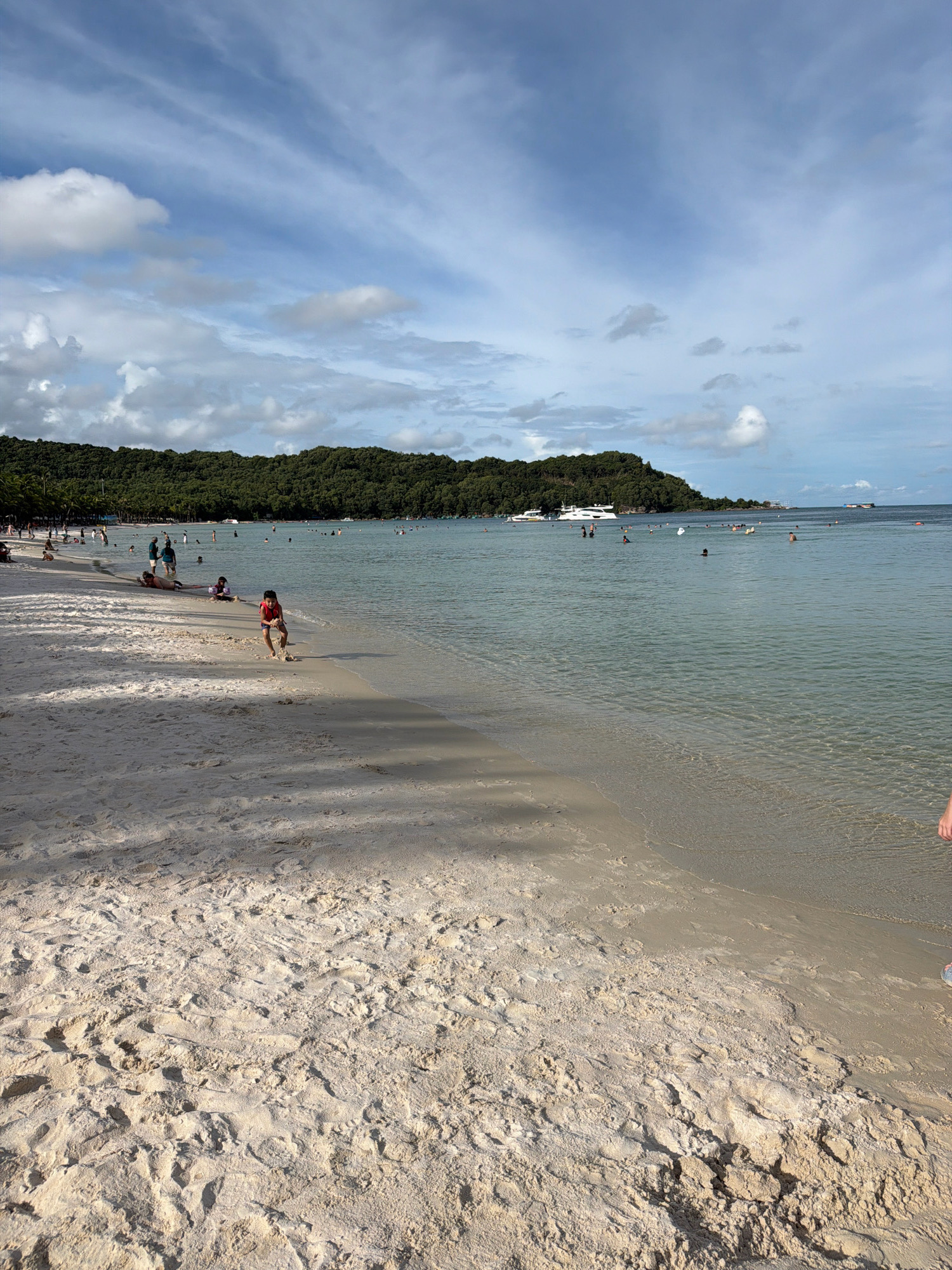 This is a photograph of the beach at Long Beach (Bãi Trường) in Phú Quốc, Vietnam. The scene depicts a wide, sandy beach in the foreground with footprints and shadows cast by palm trees. In the middle ground, the shallow, clear turquoise water laps at the shore. Numerous people are visible in the water, swimming and wading. Further out, several boats, including a large white yacht, are anchored or cruising. The background features a dense, green, tree-covered island or peninsula. The sky above is a mix of blue sky and scattered white clouds, with streaks of cirrus clouds visible. The image captures a typical day at a popular beach resort.