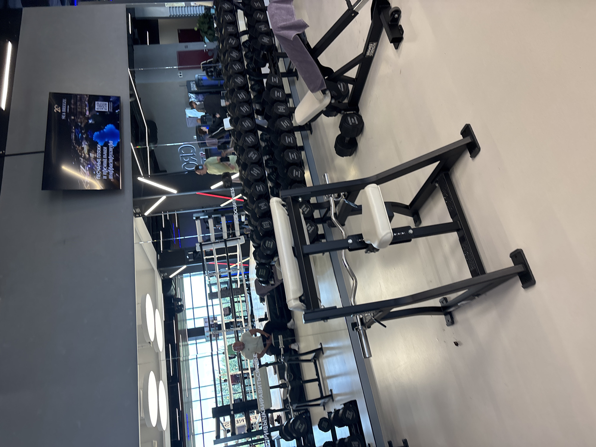 The image shows the interior of a modern, well-lit gymnasium. In the foreground, a weight bench with a padded seat and backrest is visible, with two black dumbbells on the floor beneath it. To the left, a squat rack or bench press rack is partially visible. Behind these machines, a large mirror reflects the scene, showing rows of dumbbells neatly arranged on racks.
In the midground, through the mirror's reflection, two individuals are visible. One person, wearing a light-colored t-shirt and dark pants, is sitting on a bench near a boxing ring. Another person, in a light green t-shirt, is standing. The boxing ring itself is visible in the background, with ropes and corner posts.
Further in the background, more gym equipment, including weight machines and what appear to be more boxing ring structures, can be seen. A large screen mounted on the wall displays an advertisement for "SEA BREEZE," featuring images of beaches and text in Russian. The flooring appears to be a light-colored, polished material, and the ceiling features modern lighting fixtures.
The gymnasium is designed with a sleek, contemporary aesthetic, utilizing a neutral color palette of gray, black, and white, with touches of blue in the lighting and on the advertising screen. The presence of a boxing ring suggests this facility caters to various fitness disciplines, including combat sports. The overall impression is one of a clean, organized, and well-equipped fitness center. The image does not provide specific details that would allow for pinpointing a location within the city of Monolit, Russia.