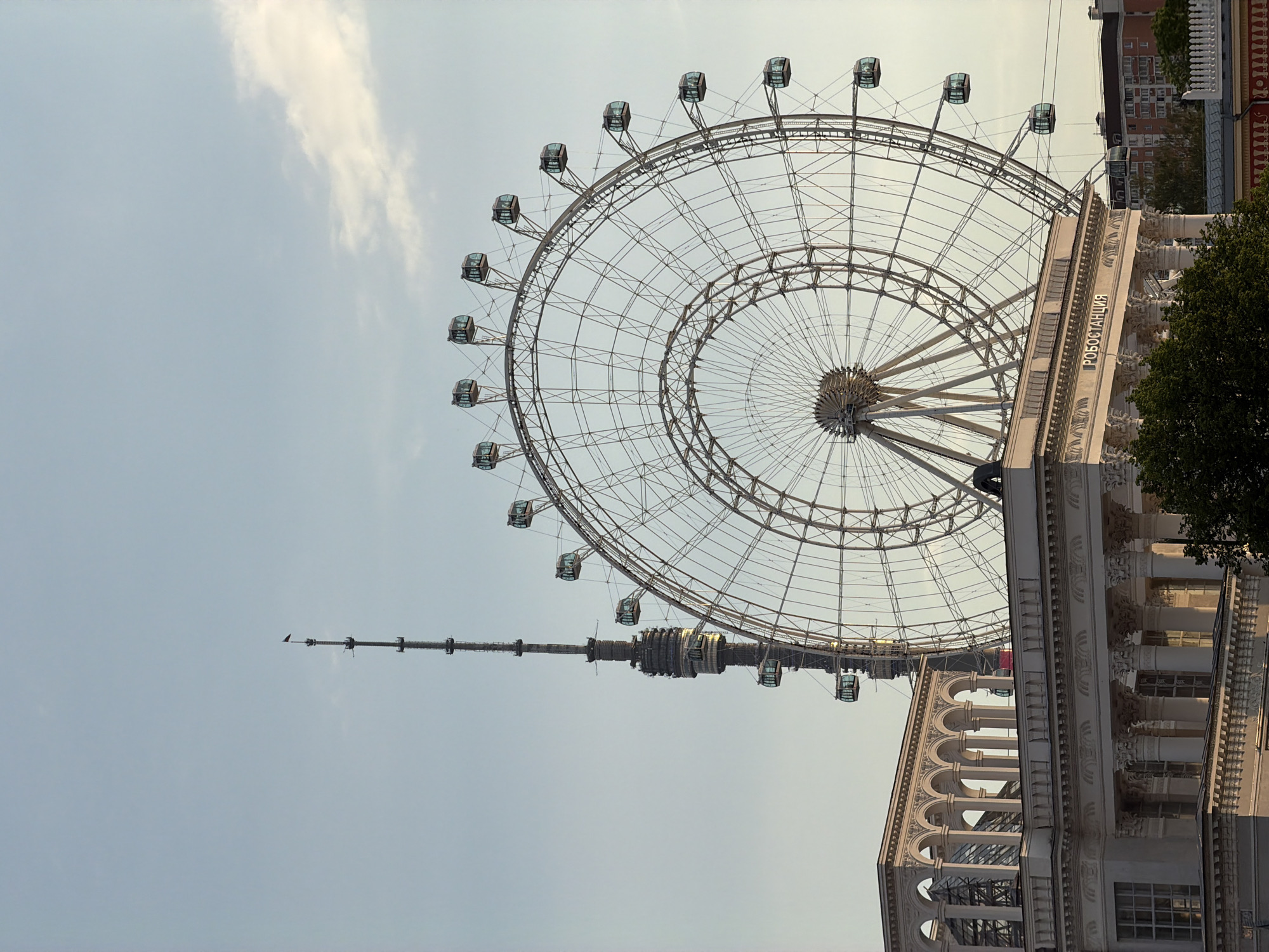 The image depicts a Ferris wheel and a structure resembling a radio tower in Moscow, Russia. The Ferris wheel is a large, silver-colored structure with enclosed gondolas, suggesting a modern design. A building with classical architectural features is also visible, positioned to the right of the Ferris wheel. The sky is pale blue with faint clouds. There are no people visible in the image. The location is likely within the VDNH park, where both the Ferris wheel and the Ostankino Tower are situated.