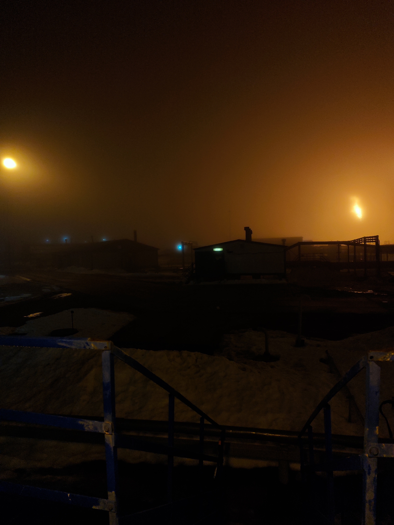 The image shows a foggy night scene in what appears to be an industrial or outpost area of Unknown, Russia. In the foreground, there's a section of a blue metal railing or fence. Beyond the railing, the scene is dominated by a heavy fog, lit by yellow or orange streetlights. Several dark buildings are visible in the middle distance, and there's a structure that looks like an open framework or scaffolding on the right. There are patches of snow on the ground. The overall impression is one of isolation and sparse activity in a cold, foggy environment.