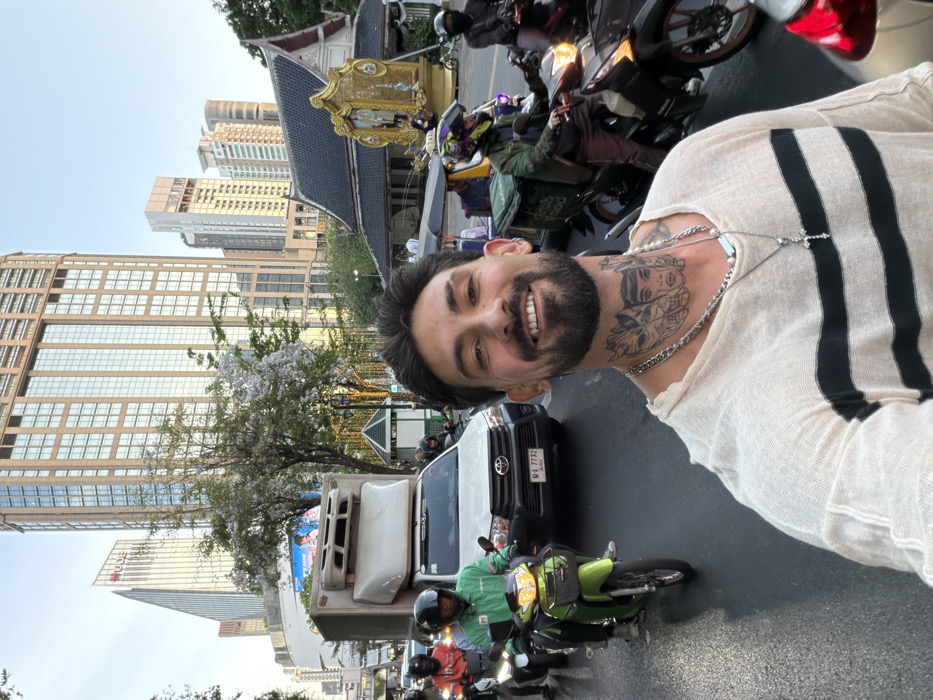 The image captures a bustling street scene in Bangkok, Thailand, specifically near the Ratchaprasong intersection, with the Erawan Shrine prominently visible on the right.
In the foreground, a male individual is centered, smiling directly at the camera. He has dark hair, a full beard, and numerous stylized face tattoos covering his throat and upper chest. He wears a loose-fitting, light-colored, long-sleeve top with two dark horizontal stripes across the chest and two silver necklaces, one featuring a cross pendant.
Behind him, a busy urban street is congested with traffic. A white Toyota pickup truck with a large, white refrigerated unit or cargo box on its bed is visible, bearing the Thai license plate "พฉ 7732." To the left of the truck, a motorcyclist wearing a green uniform and helmet operates a lime-green scooter. Several other motorcycles are present, including a delivery rider with a green "dmb food" insulated bag on their bike, positioned to the right of the main subject. A yellow tuk-tuk is partially visible behind other vehicles on the right. All visible motorcyclists wear helmets.
The background displays a dense cityscape dominated by modern skyscrapers with glass facades, including the distinctive UOB building with its angled roof on the far left. On the right, the Erawan Shrine, a traditional Thai structure with a dark, multi-tiered roof and an elaborate golden statue, stands amidst the modern architecture. Trees with light purple blossoms are visible, indicating foliage along the street. The sky is clear and bright, suggesting daytime conditions.