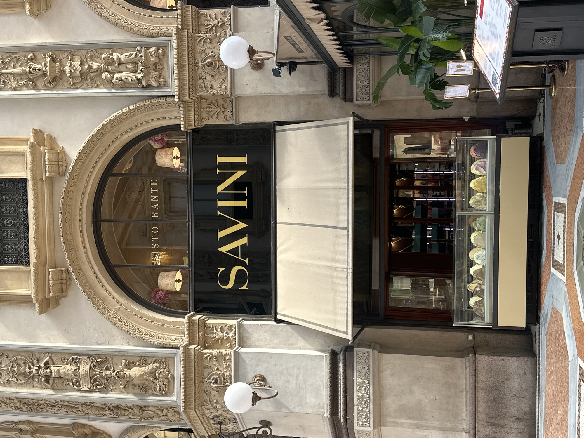 The image shows the exterior of Savini, a historic cafe and restaurant located in the Galleria Vittorio Emanuele II in Milan, Italy. The establishment features a large arched window above the entrance, with ornate architectural details surrounding it. A black sign with gold lettering displays the name "SAVINI." Below the sign, a beige awning covers the entrance to the cafe. In front of the entrance, a refrigerated display case showcases various flavors of gelato. The surrounding architecture is characterized by classical columns, intricate carvings, and decorative lamps. The floor is paved with a mosaic of colored marble tiles. The scene is brightly lit, suggesting it is daytime. There are no people clearly visible in the image, but reflections in the windows hint at the interior.