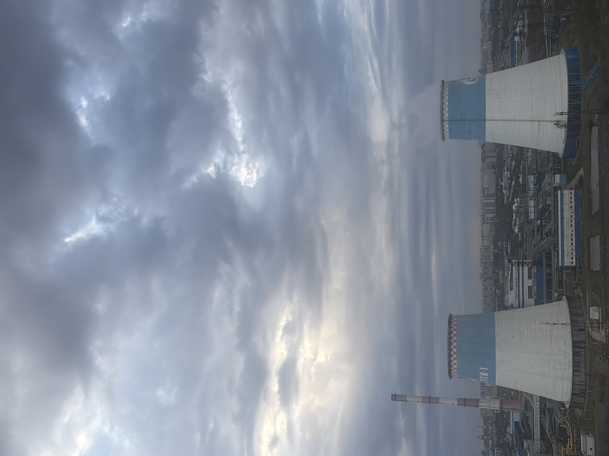 The image depicts an industrial landscape, likely a thermal power plant, under a heavily overcast sky. The scene is dominated by two large cooling towers and a tall chimney, with a sprawling industrial complex and distant city buildings visible in the background.
In the foreground and midground, two massive hyperboloid cooling towers stand out. The one on the right side of the frame is primarily white with a wide blue band around its upper section. Its very top rim features a checkered pattern of white and a reddish-brown or orange color, and steam or vapor is visibly rising from its opening, indicating active operation.
To the left, another cooling tower shares a similar white and blue design, but its upper rim is adorned with a more distinct red and white checkered pattern. Below the blue band, clearly visible on the white surface of this tower, is the Cyrillic text "ТЭЦ-19". "ТЭЦ" stands for "Теплоэлектроцентраль" (Thermal Power Plant) in Russian, identifying the facility. Adjacent to this cooling tower is a tall, slender chimney with alternating red and white stripes.
The sky above is filled with thick, gray clouds, varying in density and shading. While generally overcast, there are brighter patches where light penetrates, suggesting it is daytime, possibly under heavy cloud cover or during a specific time like late afternoon or early morning. No direct sunlight is visible. The overall weather appears cool and possibly gloomy due to the dense cloud cover.
The background reveals a complex network of industrial structures, pipes, and buildings, gradually fading into a hazy cityscape of distant urban dwellings and infrastructure. No people are visible in the scene. The location is Saint Petersburg, Russia.