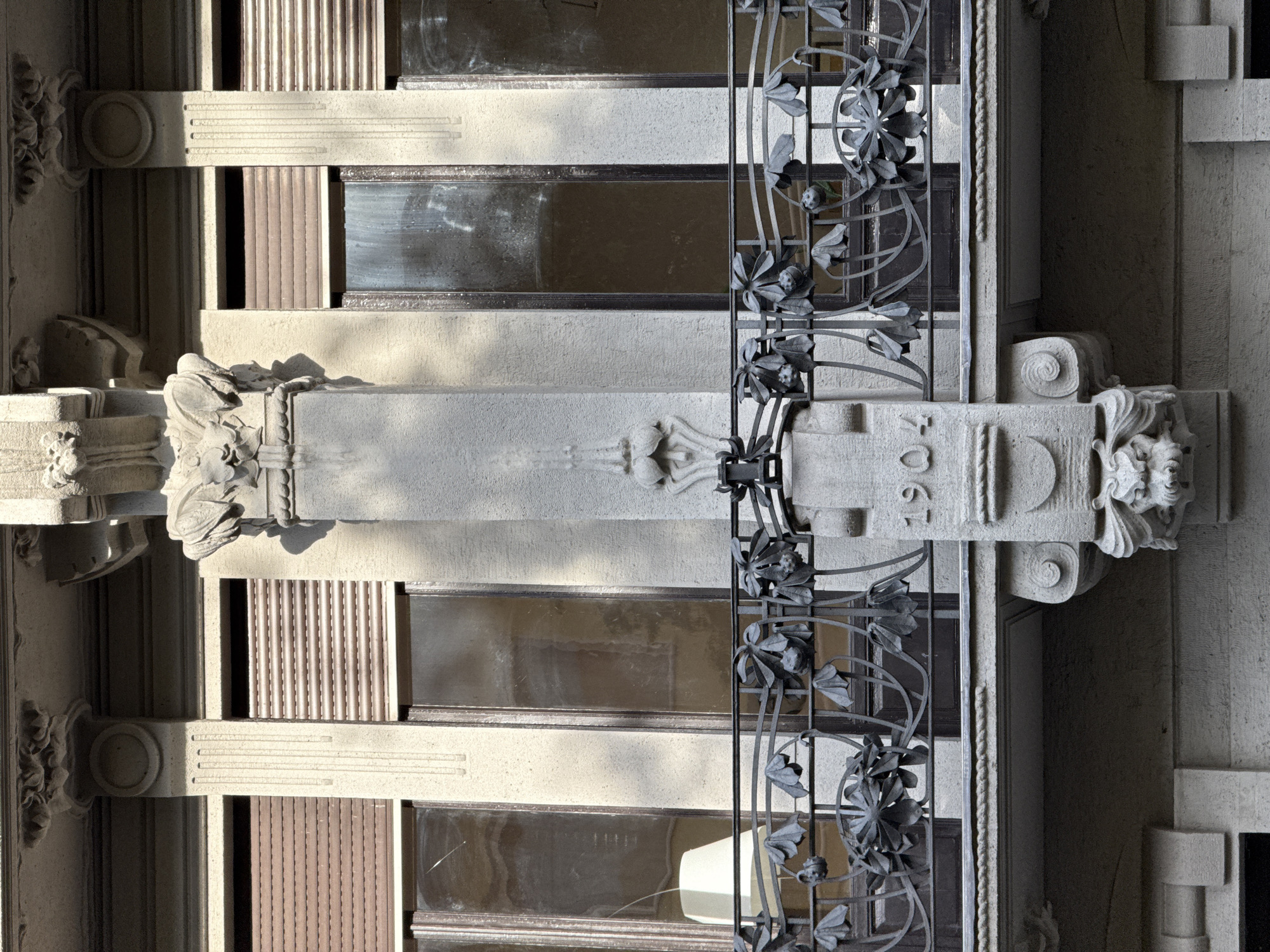 A detailed view of a building facade in Milan, Italy, featuring a decorative iron balcony with floral designs. The facade is characterized by horizontal layers of textured stone, separated by rectangular windows. The balcony's ironwork is intricate, with large floral motifs repeating along its length. A vertical stone pillar with ornate carvings divides the balcony and windows. "1906" is inscribed on a part of the stone structure. The building's architecture shows detailed craftsmanship and the use of stone.