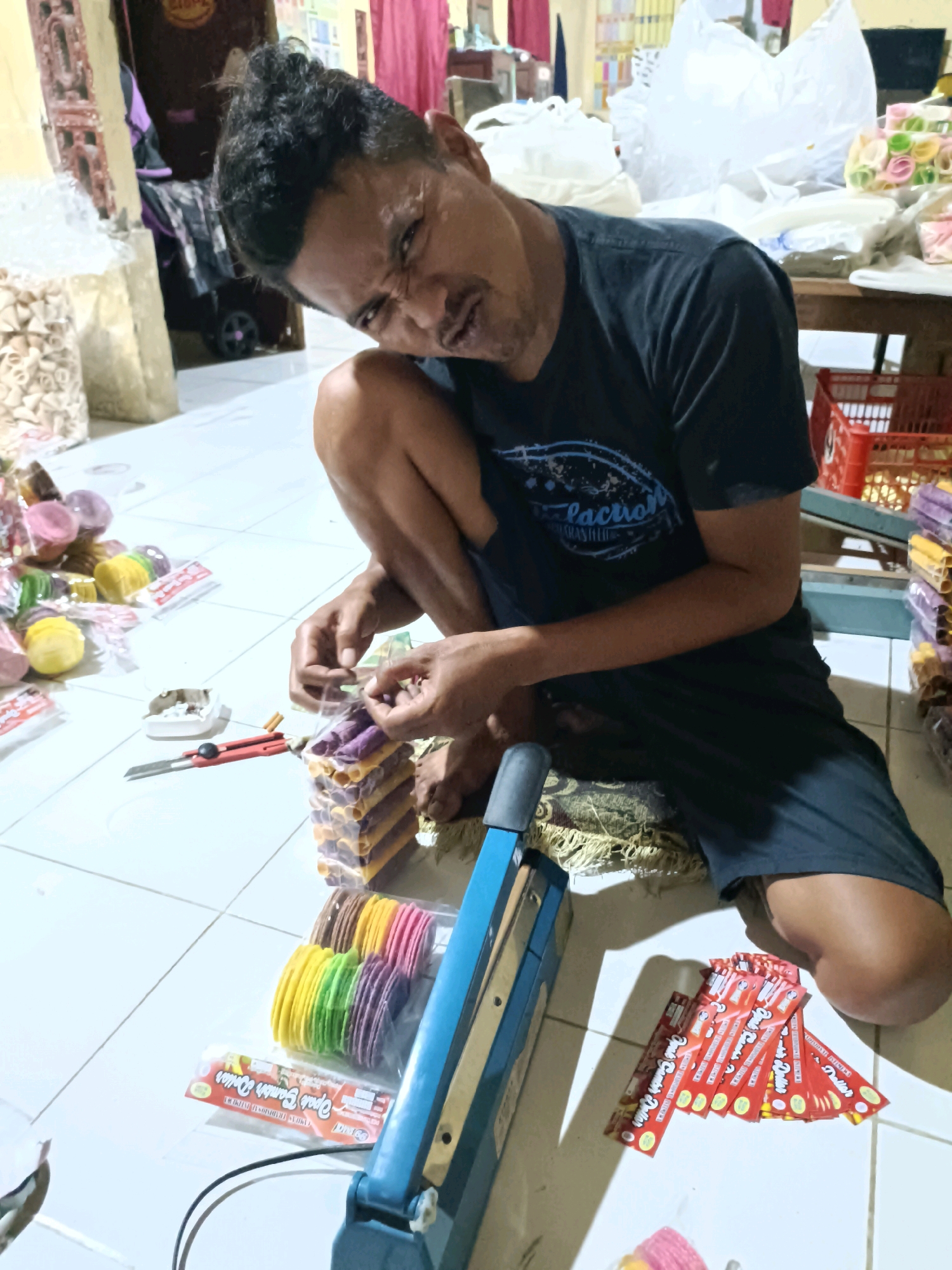 In Ploso, Indonesia, a man is seen crouching on a tiled floor in what appears to be a home or workshop. He is actively engaged in packaging colorful snacks. His focus is directed downwards, and his facial expression suggests concentration. In his hands, he is manipulating plastic wrap around stacks of the snacks, which are cylindrical and appear to be of various colors such as purple, yellow, and brown.
To his right, a blue heat-sealing machine is positioned, indicating the process of sealing the snack packages. Nearby, a red utility knife and an ashtray with cigarette butts are visible on the floor. Scattered around the area are more packaged snacks, some in larger plastic bags and others individually wrapped. The background reveals a cluttered environment with stacked items, plastic bags, and what looks like shelving or furniture. A red plastic basket is also present. The overall scene suggests a small-scale production or packaging operation of snacks.