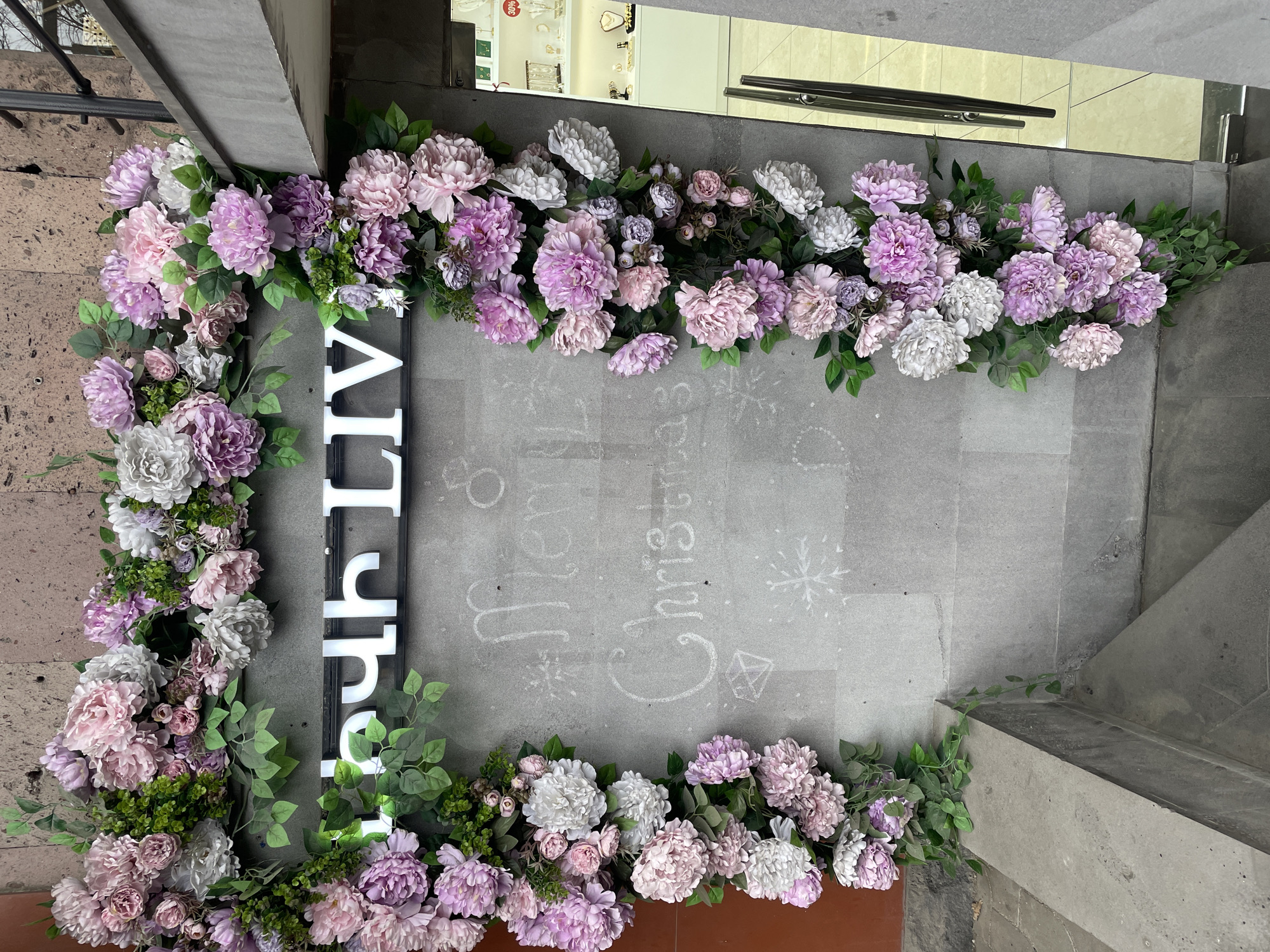 The image presents the exterior entrance of Kari LIV Jewelry, located at Abovyan Street 12 in Yerevan, Armenia. A decorative floral garland, consisting of artificial flowers in various shades of pink, purple, and white, intertwined with green foliage, adorns the upper perimeter and right side of the entrance. Above the floral arrangement, an illuminated sign displays "ԿԱՐԻ LIV" in prominent white, capitalized letters on a dark rectangular frame. The grey, textured stone wall beneath the sign and to the left of the glass door features handwritten chalk inscriptions that read "Merry Christmas," accompanied by drawings of a star, two diamond shapes, and a snowflake. To the right, a reflective glass door with a metallic vertical handle offers a partial view of the brightly lit interior, revealing white display cases holding various jewelry items. A small red "30%" discount sign is visible within the reflection. On the left side of the frame, grey stone steps lead downwards, partially obscured by the floral decoration. No individuals are present in the depicted scene.