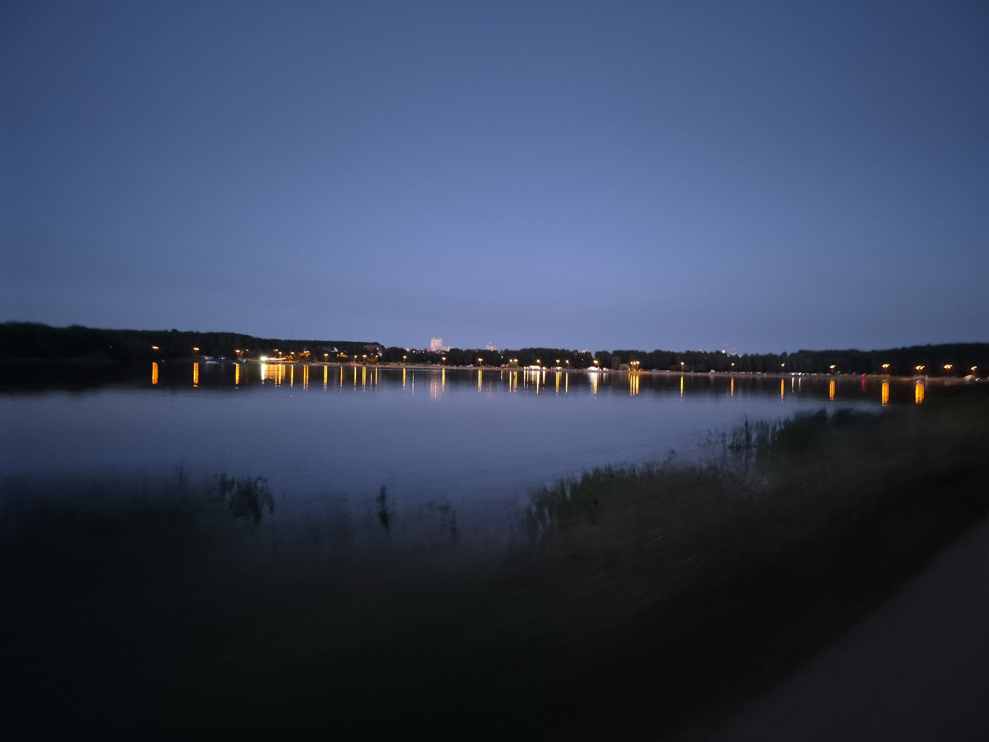The image depicts a serene twilight scene at a body of water, likely a lake or a river, in Minsk, Belarus. The sky is a deep, clear blue, transitioning to a darker hue at the horizon. On the far bank, a line of trees forms a dark silhouette against the fading light. Beyond the trees, the faint glow of city lights, including taller buildings, can be discerned.
The water itself is calm and reflective, mirroring the lights from the opposite shore. These lights appear as elongated, warm orange and yellow streaks on the water's surface, creating a beautiful, shimmering effect. Some reeds or tall grasses are visible along the near edge of the water, adding texture to the foreground. The overall impression is one of tranquility and urban beauty as night begins to fall. The specific location within Minsk is not identifiable from the image alone, but the presence of a large body of water and distant city lights suggests a park or recreational area bordering a lake or river within the city.