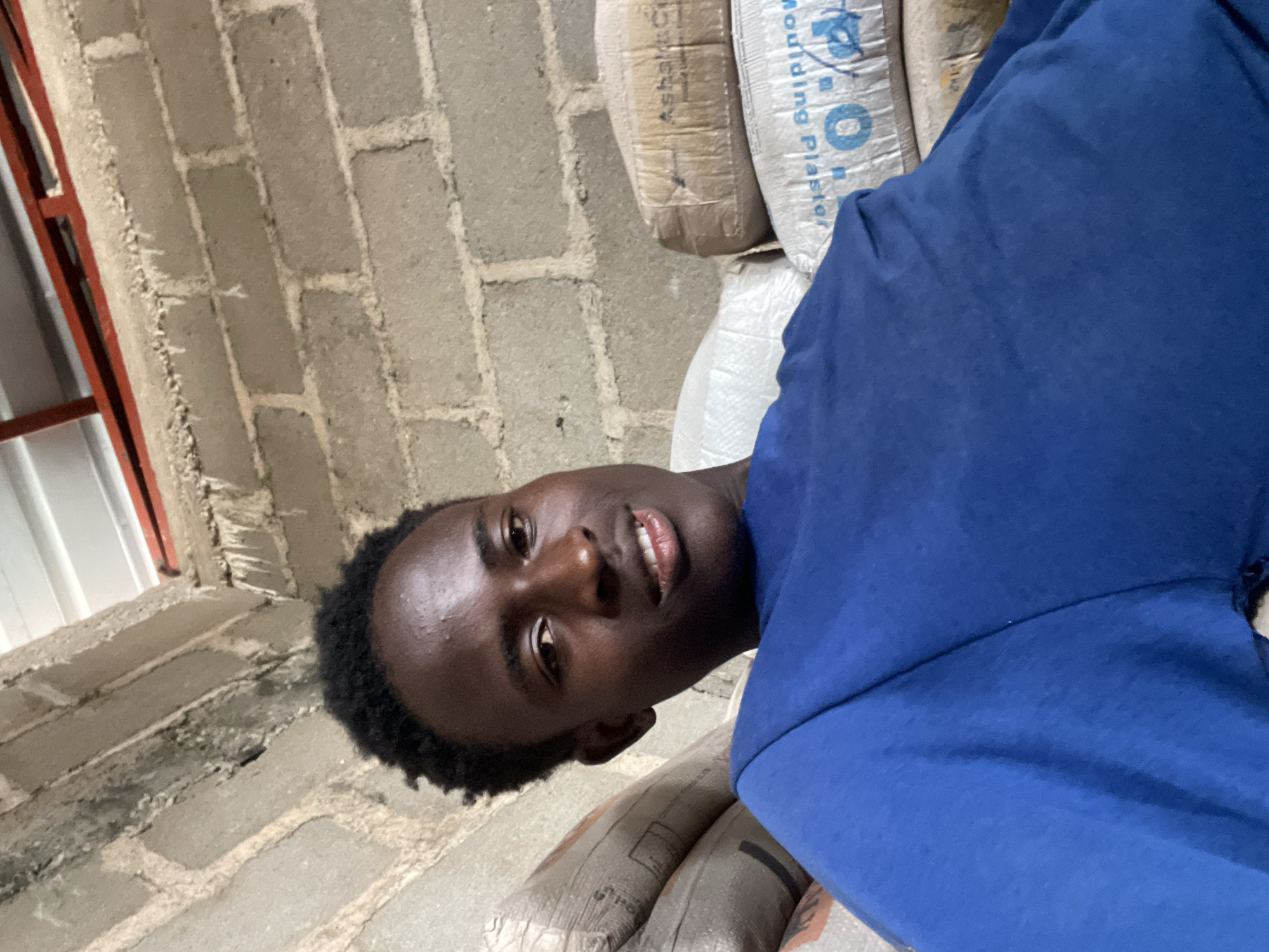 The image shows a medium shot of a young Black man wearing a blue t-shirt, taken from a slightly high angle. He has short, dark, curly hair and is looking towards the camera with a slight smile, his head tilted to his right. The background consists of a rough, grey cinder block wall and stacks of bagged materials, one with visible text that appears to be "Moulding Plaster" and a logo with the letter "O". The scene suggests an indoor setting, possibly a storage area or construction site. The lighting is natural and appears to be coming from above, casting faint shadows. The overall impression is informal, with the subject seemingly relaxed. This scene is consistent with the environment found in many parts of Jos, Nigeria, particularly in areas with ongoing construction or warehousing.