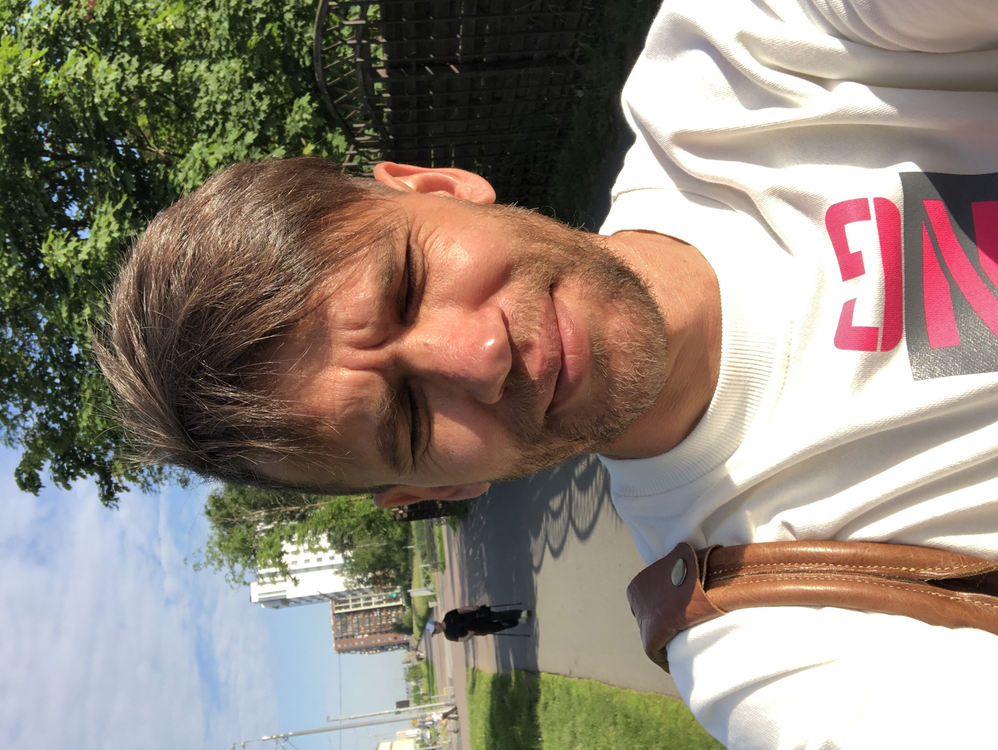 The image is a selfie taken by a man with short brown hair and a beard, wearing a white sweatshirt. He is standing outdoors on a sunny day. In the background, there is a paved walkway, green grass, trees, a black metal fence, and modern buildings. A person in the middle ground is walking on the path with trekking poles. The setting appears to be a residential area in Saint Petersburg, Russia, possibly during a walk in a park or along a street with apartment buildings. The man's facial expression is neutral, with his eyes closed or squinting due to the sunlight.