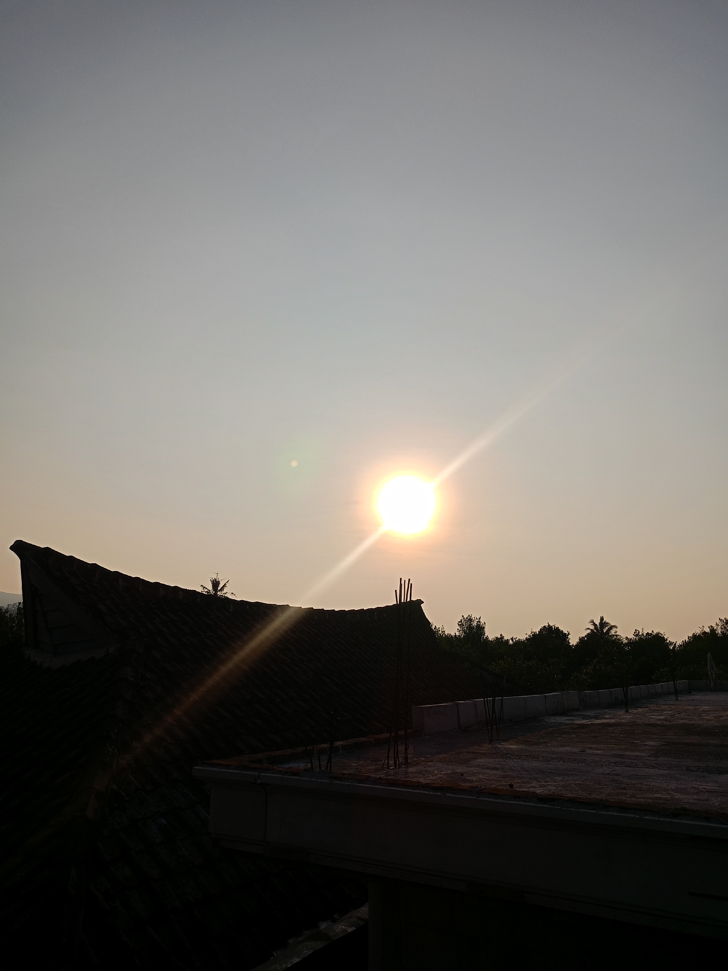The image depicts a silhouetted view of rooftops against a clear sky during either sunrise or sunset. The sun, a bright orb, is positioned in the upper center of the frame, emitting a lens flare and sunbeams that diagonally cross the sky. In the foreground, the dark silhouette of a tiled roof with a distinctive curved edge dominates the left side of the image. To the right, a flat rooftop is visible, showing exposed rebar and a low concrete barrier. Beyond the rooftops, a line of trees, including a palm tree, creates a dark, dense foliage against the horizon. The overall scene suggests an outdoor environment in a location that may be a residential or urban area, given the presence of rooftops and construction elements. The lighting indicates a time of day when the sun is low in the sky.