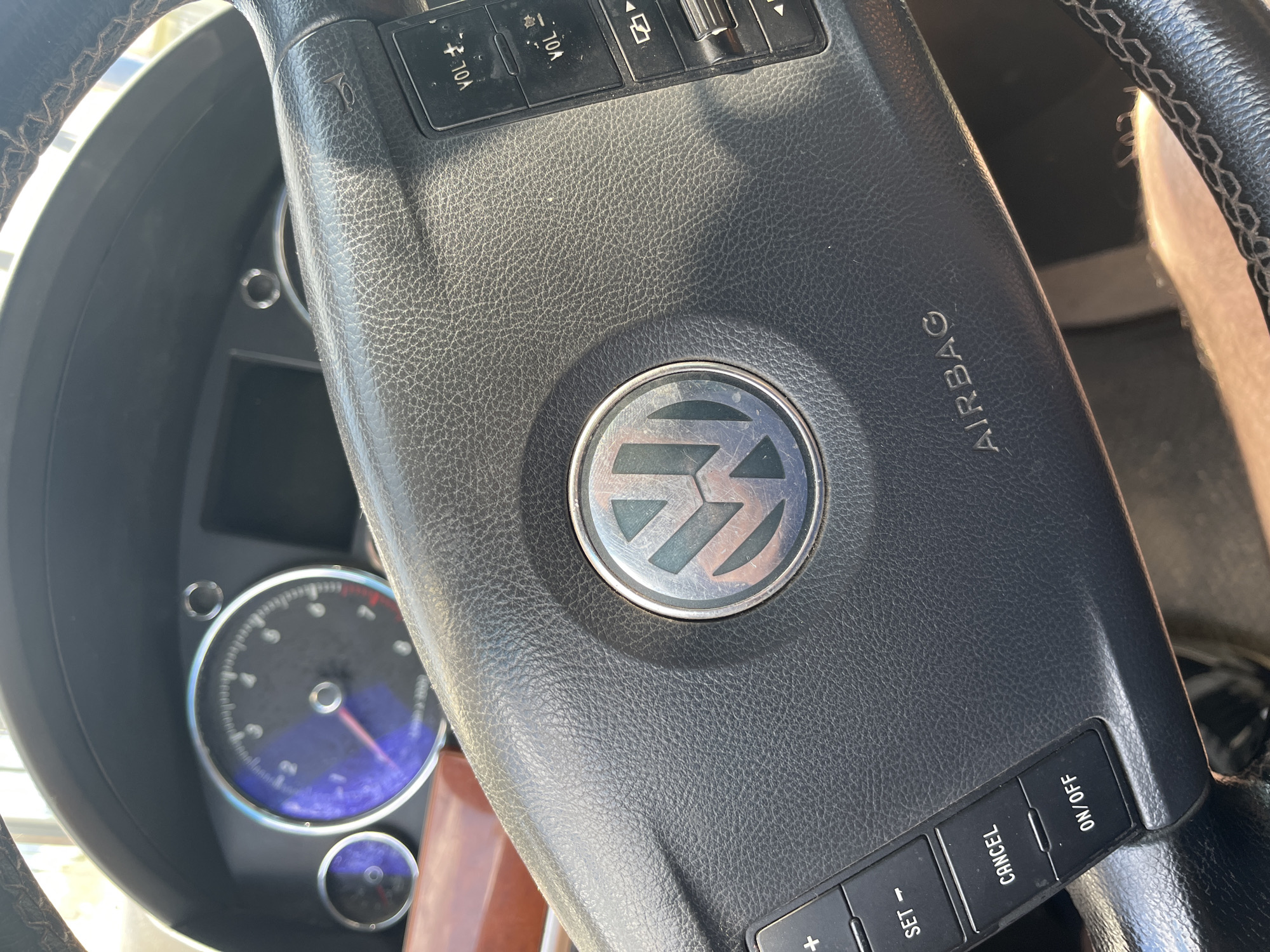 The image shows a close-up view of the steering wheel and dashboard of a car. The steering wheel is black leather with stitching and features several buttons. A prominent circular emblem with the Volkswagen logo is centered on the airbag cover. Text indicating "AIRBAG" is visible on the steering wheel. To the left of the steering wheel, parts of the car's instrument cluster are visible, including a speedometer with a red needle pointing towards a blue shaded area. Below the speedometer is a smaller circular gauge. The scene appears to be an interior shot of a vehicle, likely parked. The lighting suggests it could be daytime. There are no people visible in the image. The location context provided is Aqköl, Kazakhstan.