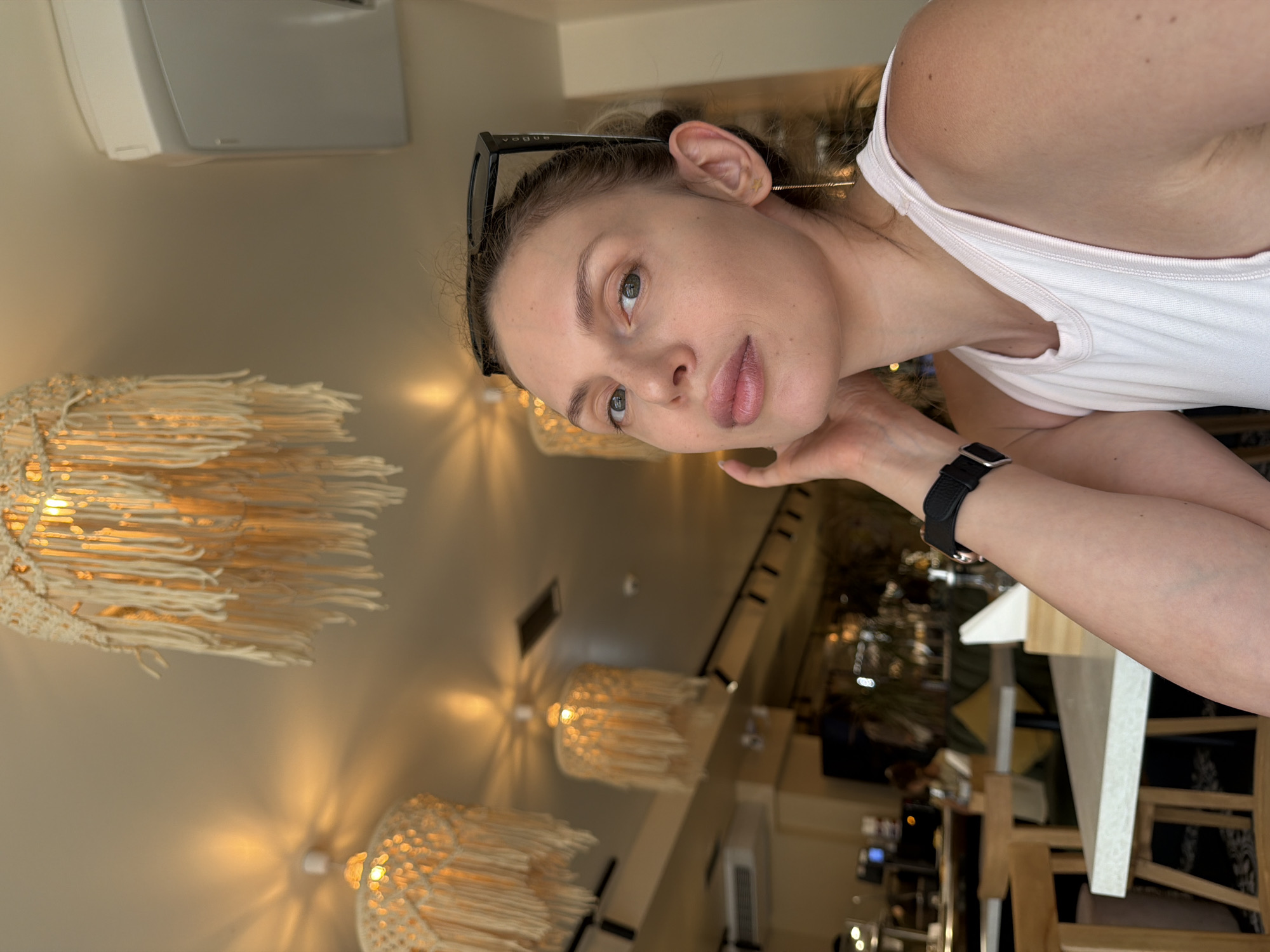 A medium shot captures a fair-skinned woman with light brown hair styled in a bun, wearing a light pink tank top and sunglasses perched on her head. She has delicate facial features, including light green eyes, a straight nose, and full lips. A thin gold earring dangles from her right ear. Her left hand is raised, touching her neck, and she wears a black wristwatch on her left wrist. The background is blurred, showing the interior of a restaurant with warm lighting and macrame lamps hanging from the ceiling. Tables and chairs are visible, along with other patrons in the distance, suggesting a relaxed atmosphere. The overall impression is that of a casual snapshot taken in a cafe or restaurant in Budva, Montenegro, during daylight hours.