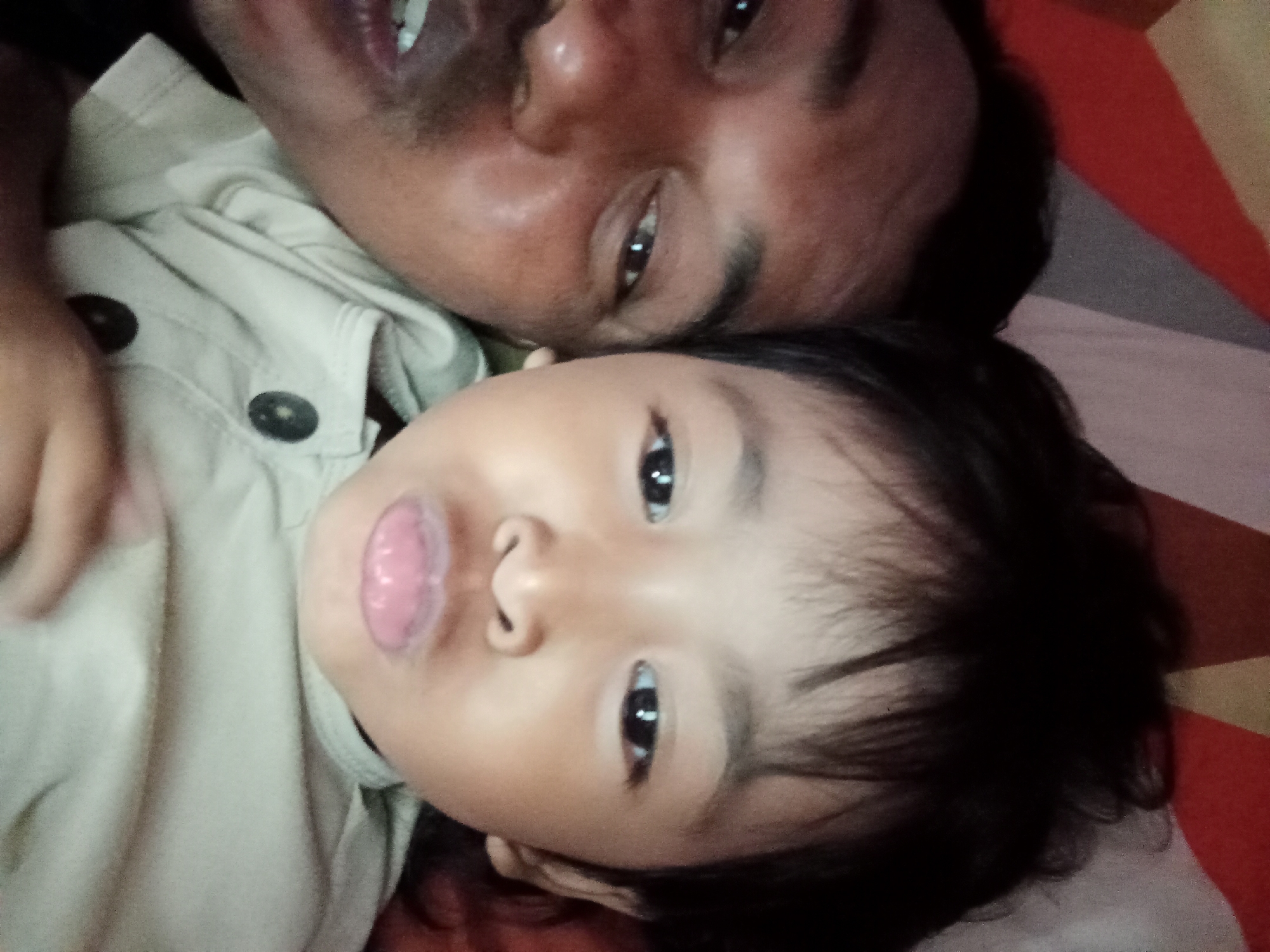 The image is a close-up, selfie-style photograph of two people, likely an adult and a child, lying down. The adult, presumably a man based on facial hair and bone structure, is positioned in the upper part of the frame. He has his eyes open and appears to be smiling slightly, with his mouth partially open showing teeth. The child is positioned below the adult, with their face closer to the camera. The child's eyes are open, and their tongue is sticking out, which gives a playful or curious expression. Both individuals are wearing light-colored clothing, possibly a collared shirt, with visible buttons. The background shows a textured surface, possibly a bed or a mat, with red and beige striped patterns. The lighting suggests it might be indoors, possibly during the day given the brightness. The overall mood appears affectionate and casual, capturing a moment of closeness between the two subjects. There is no visible text in the image.