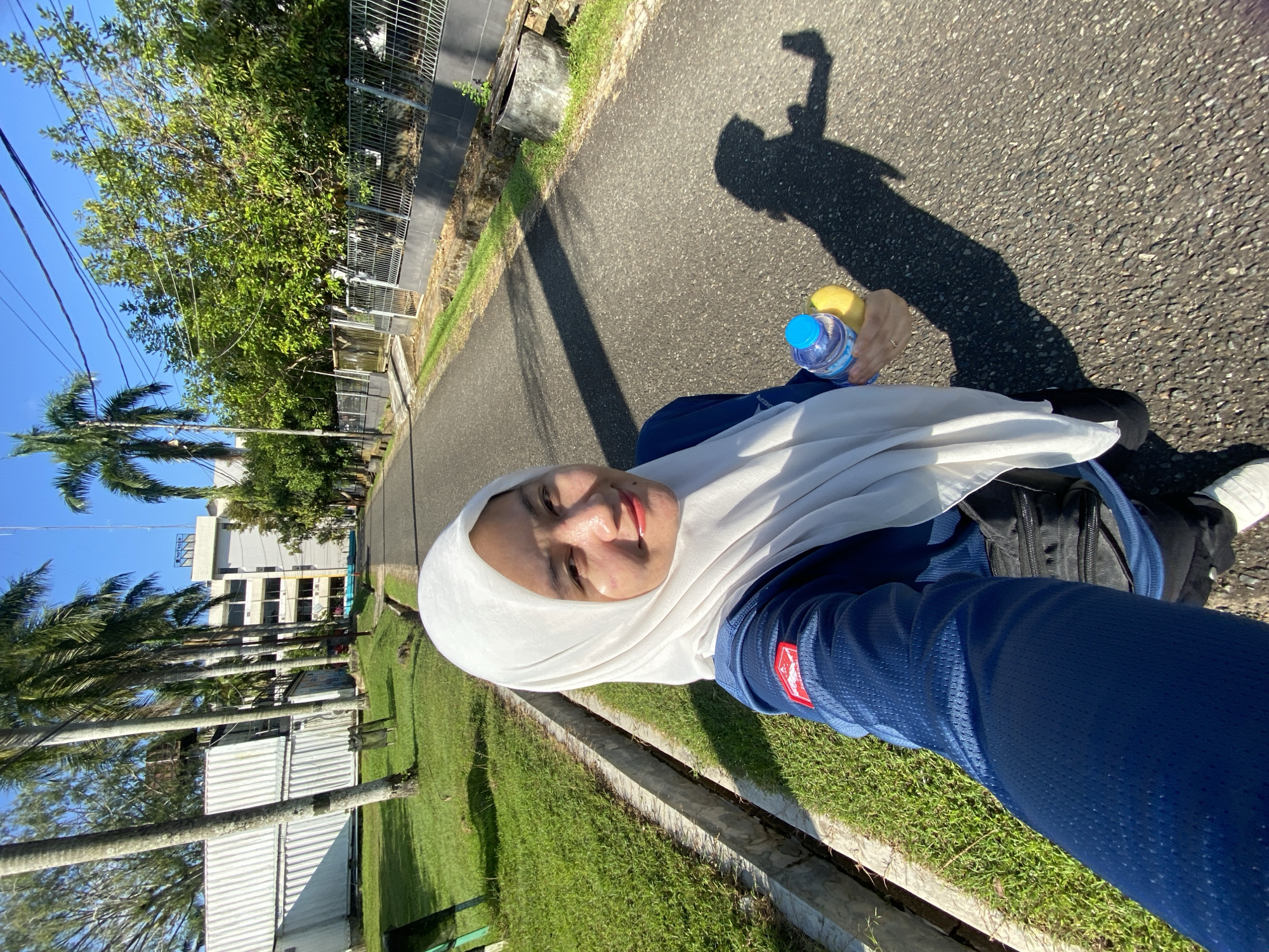 A woman wearing a white hijab and a blue long-sleeved shirt is taking a selfie outdoors. She is holding a bottle of water and a yellow object, possibly a fruit. The background shows a paved road lined with green grass, palm trees, and buildings, suggesting an urban or campus setting. The woman is casting a shadow on the road, indicating a sunny day. The image appears to have been taken in Banjarbaru, Indonesia, given the presence of tropical vegetation.