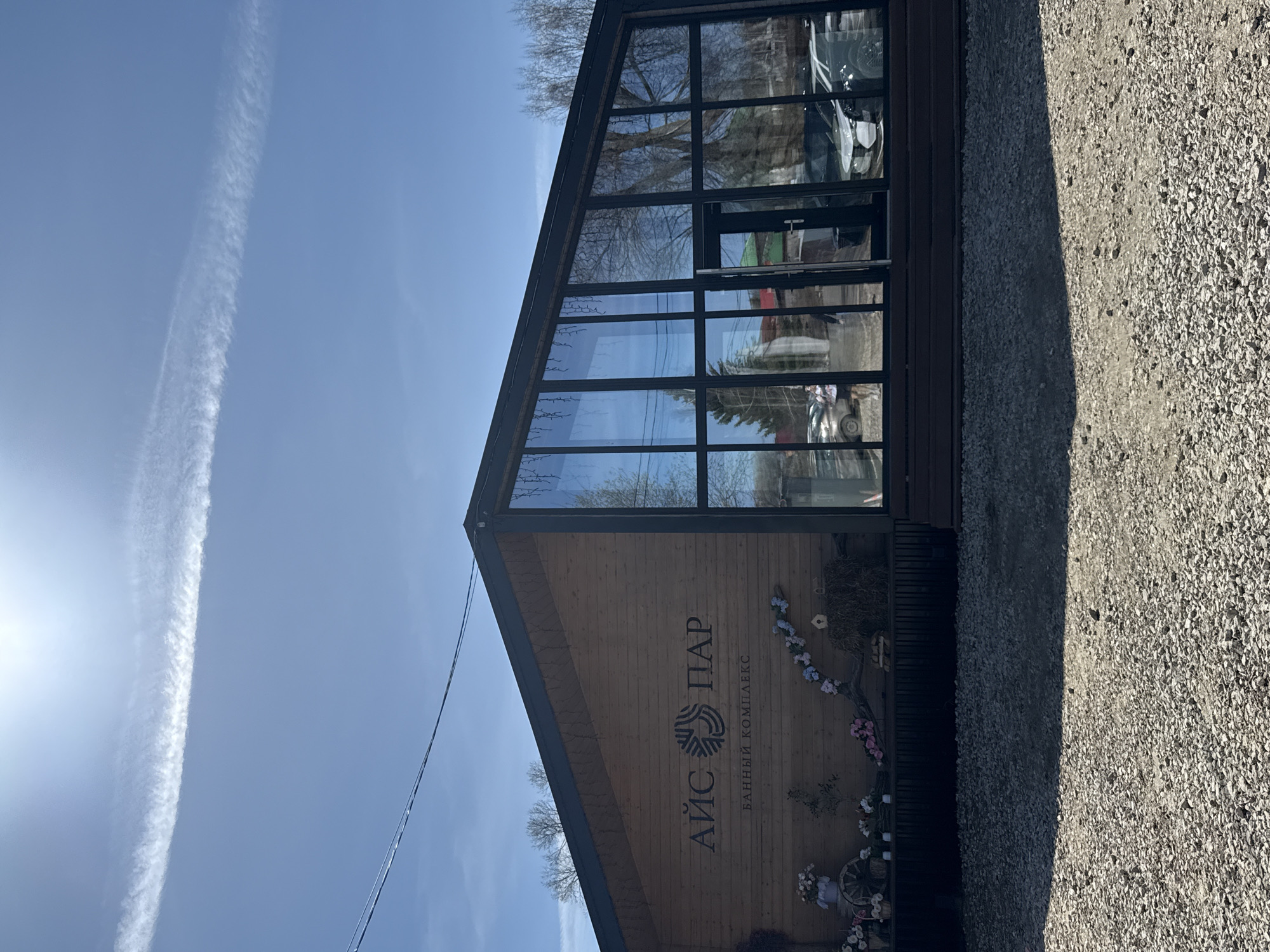 Here's a description of the image:
The image shows the "Ice Park" bathing complex located in Kazan, Russia. The building features a triangular architectural design with a facade of wooden planks. The building also has a large window frame consisting of rectangular panels. The sky is blue with a visible contrail. There is gravel surrounding the building.