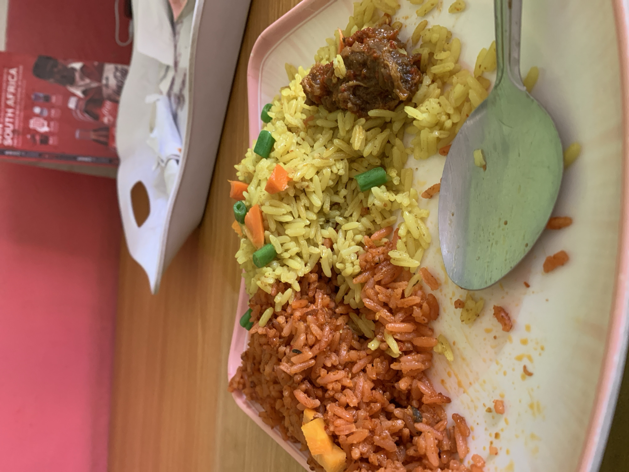 Within an indoor setting in New Stadium, Nigeria, a meal is presented on a light pink rectangular plate with a raised rim. The plate contains two distinct preparations of rice: a substantial portion of orange-red rice occupies the left side, interspersed with yellow solid pieces; to its right, a smaller pile of yellow rice is visible, mixed with green bean segments and orange carrot pieces. A dark, reddish-brown portion of cooked meat, possibly stewed, rests on the yellow rice. A metallic spoon lies on the lower right of the plate, partially covered by scattered rice grains and residue, indicating consumption. The plate itself exhibits marks of previous use, with scattered food particles. The meal is served on a light brown wooden table. In the midground, a white rectangular serving tray with cutout handles contains crumpled white paper or napkins. The background features a solid vibrant pink wall on the left and a red wall or surface on the right. Leaning against the red surface is a red booklet or magazine displaying the text "SOUTH AFRICA" and "WIN" in white, alongside illustrations including Coca-Cola bottles and figures. No individuals are present in the scene.