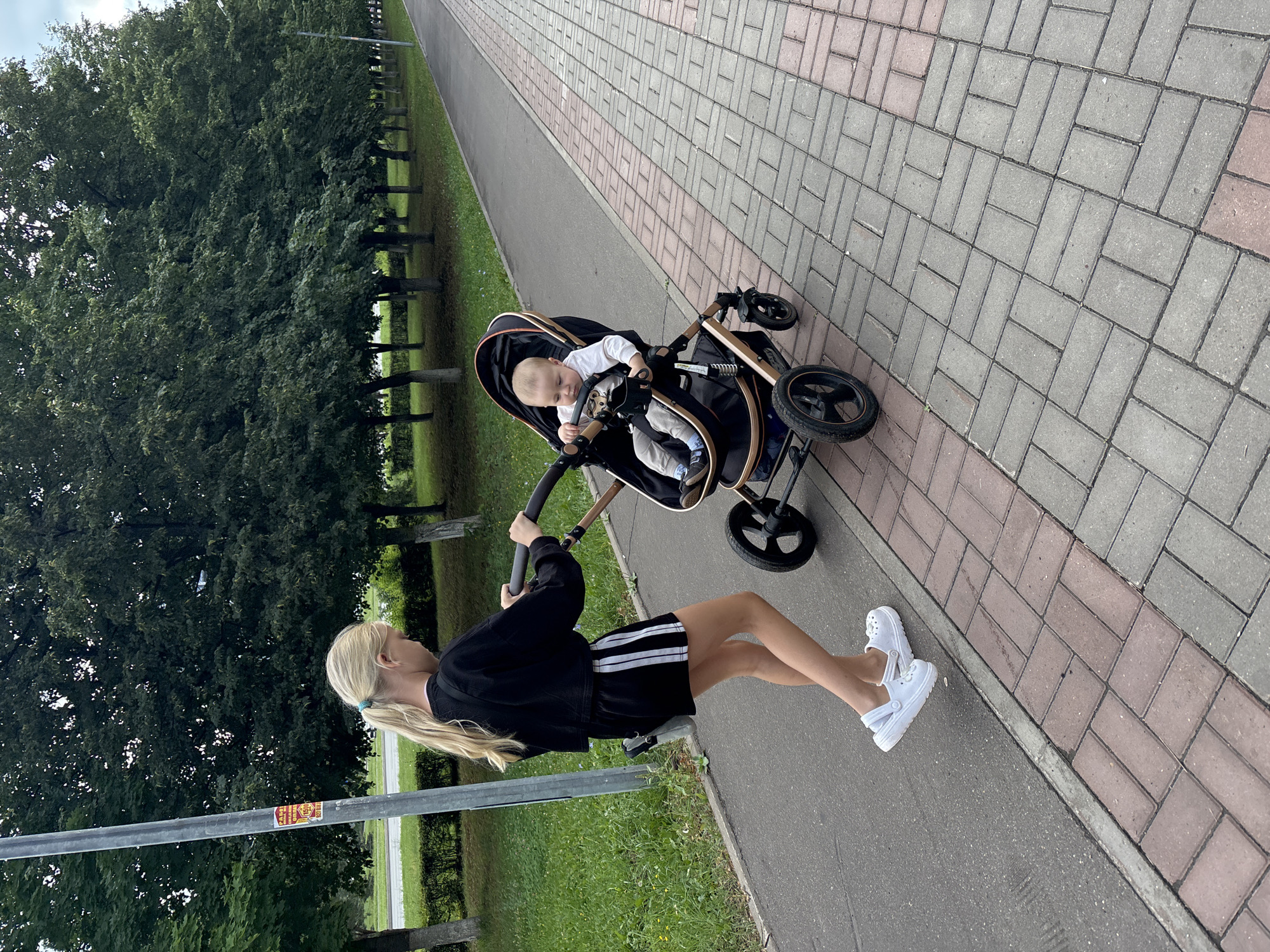 A young blonde girl with her hair in a ponytail, wearing a black cropped sweatshirt and black shorts with white stripes, is pushing a stroller with a baby in it along a paved walkway. The baby, wearing a white long-sleeved shirt and grey pants, is seated in the stroller, holding onto the handle. The walkway is made of interlocking bricks, with a section of grey bricks next to a section of red and grey bricks. To the left of the walkway is a patch of grass and a row of trees. To the right of the walkway, there is a paved road. A light pole with a sticker on it stands to the left of the girl. The setting appears to be a park or a street in Naberezhnye Chelny, Russia, given the specific details like the brickwork pattern and the presence of trees and a walkway common in such urban environments.