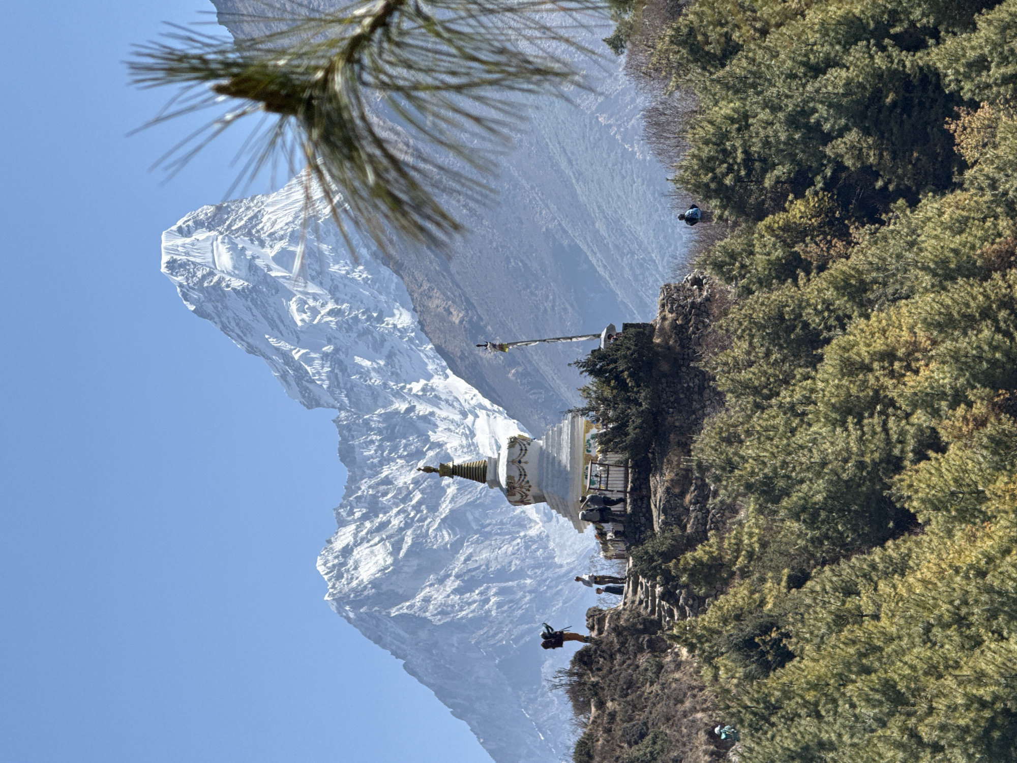 The image shows a view in Khumjung, Nepal, featuring a traditional white stupa in the middle ground. People are standing around the stupa, some appearing to be observing the landscape. Behind the stupa is a massive, snow-capped mountain towering into a clear blue sky. In the foreground, there are trees. A pine branch extends from the upper left corner of the frame, partially obscuring the view. The terrain leading up to the stupa is rocky and barren.