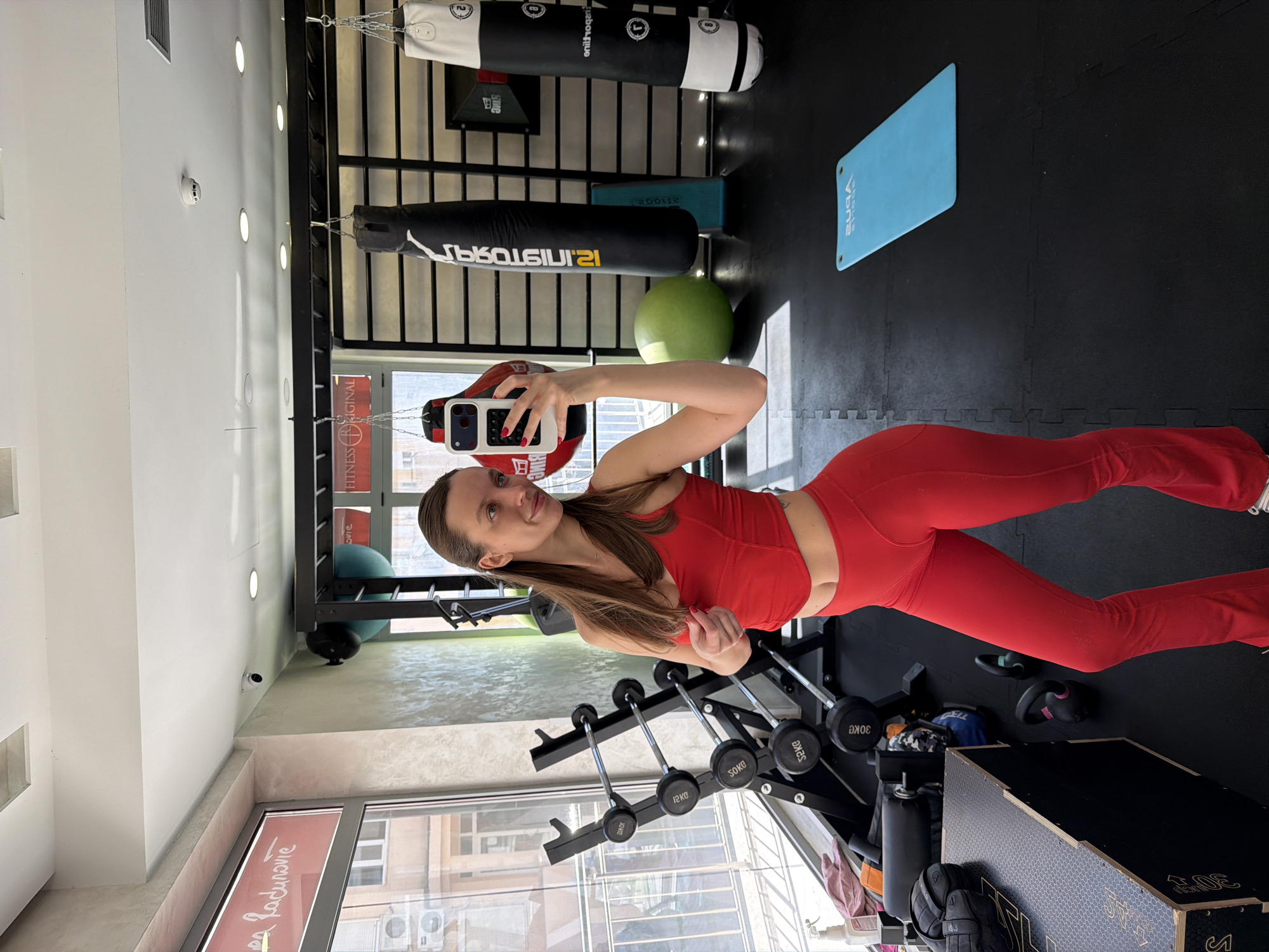 Here is a description of the image:
The image is an indoor shot of a woman in a fitness setting. She is taking a selfie, holding a smartphone with a white case and red accents, which obscures part of her face. She is wearing a red sports top and matching red leggings.
The environment appears to be a gym or fitness studio. Behind her, there's a rack with weightlifting bars and dumbbells. In the background, a window provides natural light, illuminating the space. Various pieces of exercise equipment are visible, including black punching bags hanging from the ceiling, a green exercise ball, and a blue yoga mat on the black-tiled floor.
There is lettering visible in the image but it's blurry, but the phrase "FITNESS BRIGINAL A Radunovie" can be made out. Based on this, the gym is located in the city of Budva, Montenegro.