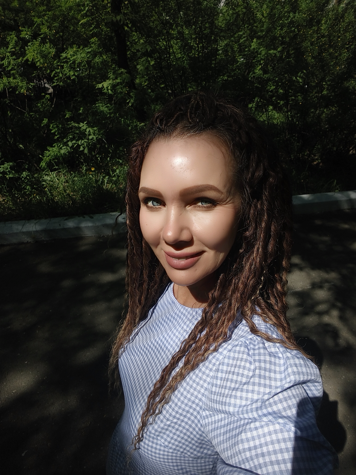 A medium close-up photograph depicts a fair-skinned woman with blue eyes, her brown hair styled in long braids, wearing a blue and white gingham-patterned blouse. The background includes a dense, green leafy area suggesting a park or garden. The presence of shadows indicates a sunny day. The location is not specified.