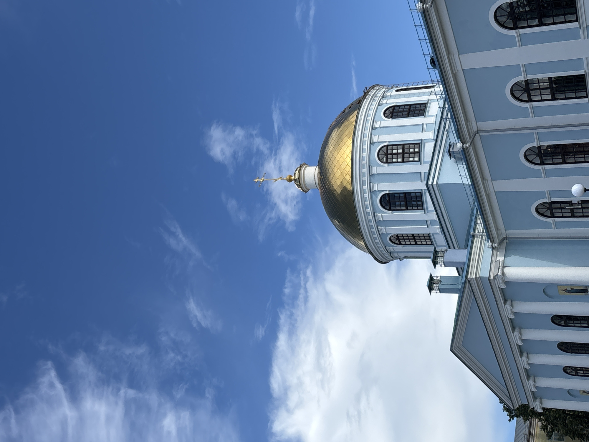 The image shows the dome and upper facade of a light blue building with white architectural details, likely a church or cathedral, in Votkinsk, Russia. The dome is golden and topped with a cross. The sky is bright blue with scattered white clouds. The building features arched windows and columns. The perspective is from below, looking up at the dome and sky.