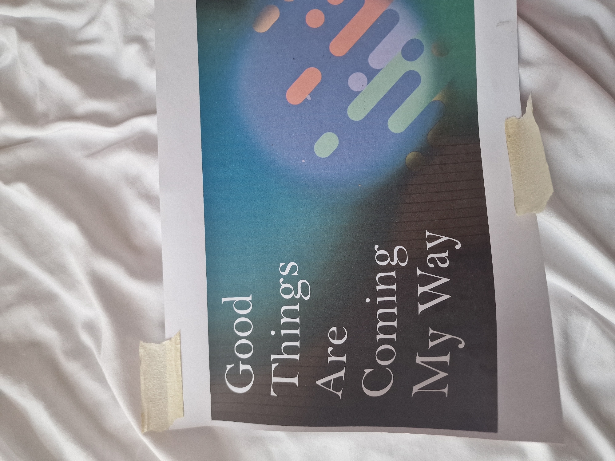 A piece of paper with the text "Good Things Are Coming My Way" printed on it is held to a white, rumpled fabric surface by two pieces of masking tape. The paper has a dark grey background with a gradient transitioning to blue and green on the right side, featuring abstract colorful shapes. The text is in a serif font and is white. The fabric surface appears to be a bedsheet. The overall scene is an indoor, domestic setting. There is no indication of a specific location in London, United Kingdom.