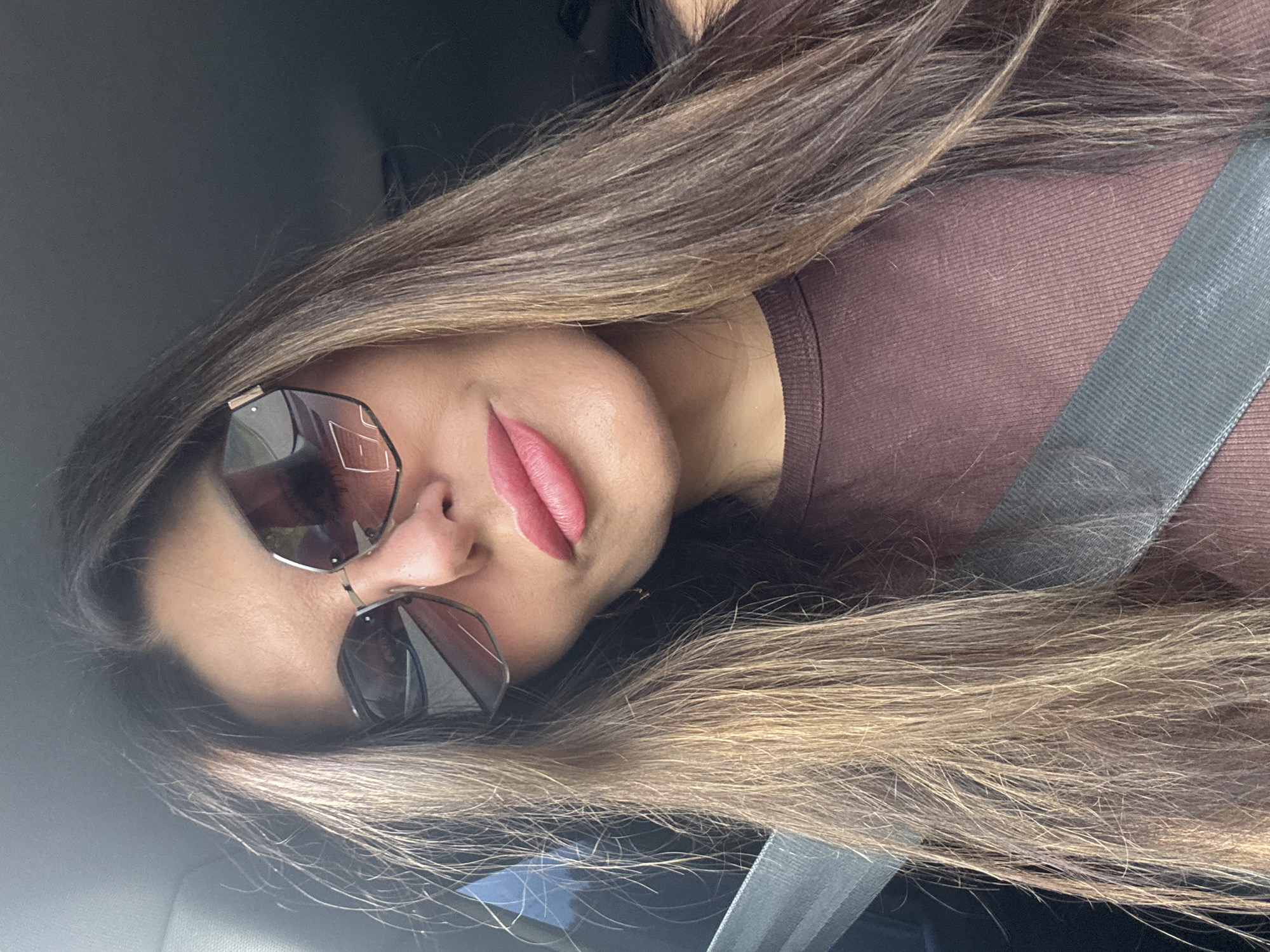 A woman wearing sunglasses and a brown shirt is in a car. Her hair is brown and falls past her shoulders. She is wearing pink lipstick. The car's seatbelt is visible across her chest. The image was taken in Chaikovsky, Russia.