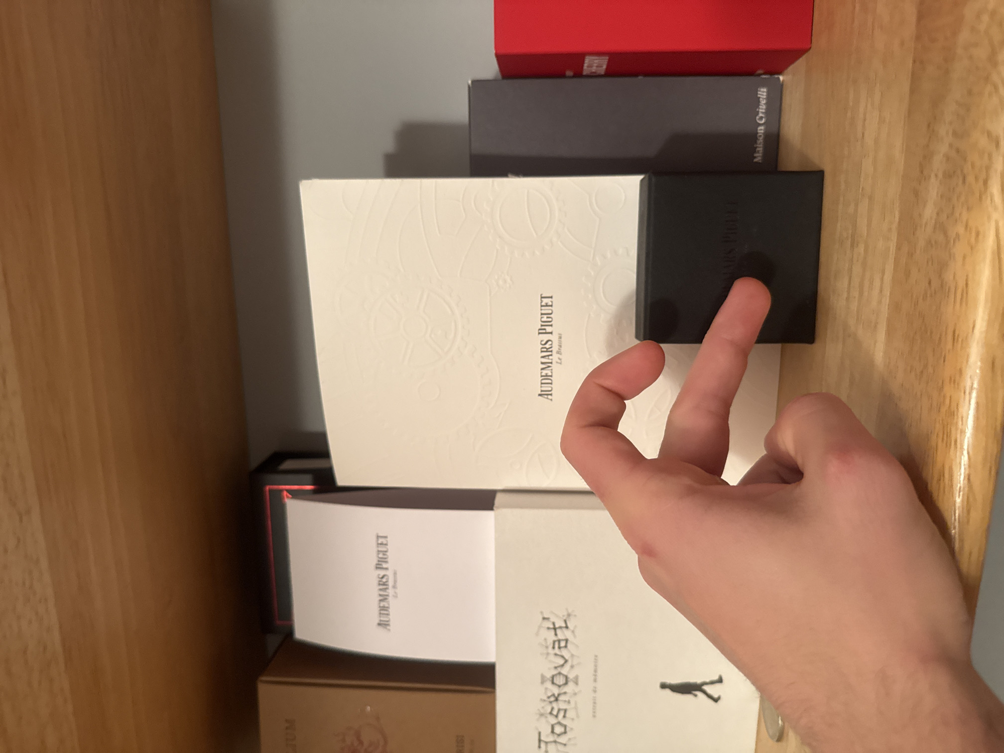 The image displays an indoor scene, possibly a shelf or cabinet, filled with various luxury brand boxes. A person's hand is visible in the foreground, pointing towards a small black box. The background includes a wooden surface on the left and a grey wall on the right.
The central subject is a collection of stacked boxes. A large white box with an embossed gear-like pattern prominently features the vertical text "AUDEMARS PIGUET Le Brassus". Adjacent to it, the hand touches a smaller black box, also labeled "AUDEMARS PIGUET". Above these, a grey box reads "Maison Crivelli" and a red box shows partial text "RISI". Below the large white box, a white open item or brochure reveals "AUDEMARS PIGUET Le Breme". Further down, another white box is labeled "Joskovat" and includes a small silhouette of a walking figure. At the very bottom, a golden box is partially visible with the text "JUM".
The activity depicted is the interaction with these boxes, possibly selecting or examining an item. The scene suggests a personal collection of high-end watches or fragrances, implying a setting of luxury goods, potentially in Milan, Italy. The lighting is uniform, characteristic of an indoor environment.