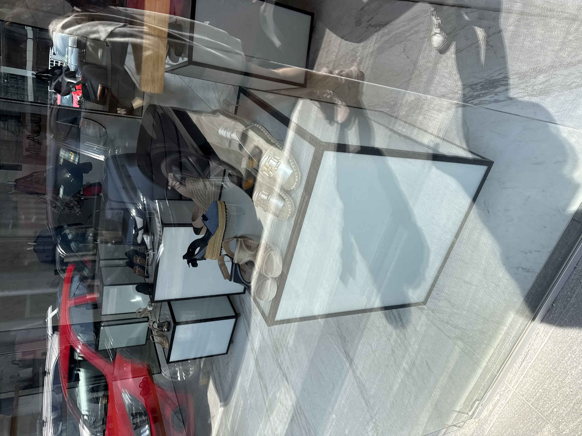 The image displays a clear daytime view through a reflective glass storefront of a retail space, identified as being located in Puebla, Mexico.
Inside the store, an array of women's footwear is presented on multiple white, rectangular display pedestals featuring dark trim. Visible shoes include gold-colored espadrille-style flats with metallic 'H' or 'T' shaped buckles, light-colored platform sandals, dark blue denim wedge sandals, nude-toned wedge sandals, black loafers, leopard-print flats, and silver high-heeled sandals. The interior floor appears to be composed of light grey or white tiles with subtle veining.
The glass facade generates significant reflections from the exterior environment. A prominent red compact sedan, possibly a Chevrolet Aveo, is reflected in the upper left portion of the image, facing left. Additional reflections include parts of other vehicles (dark and light-colored cars), indistinct building structures, and faint, reversed text such as "MIRACUL" and "MONT".
A partial reflection of a person's lower leg, wearing light-colored sneakers, along with its associated shadow, is visible in the lower right quadrant, indicating the viewpoint of the photographer. The overall lighting suggests a sunny outdoor environment. The exact street or specific district within Puebla, Mexico, cannot be determined from the visual information provided.