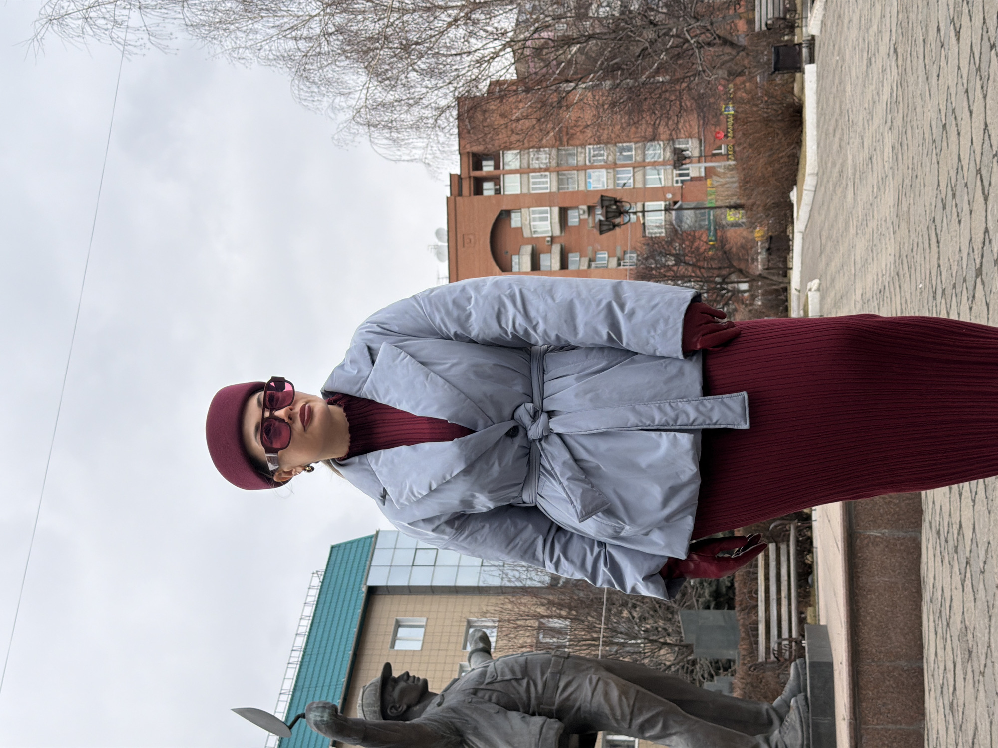 A woman stands in a city square in Krasnoyarsk, Russia, posing for a photo near a monument. She is wearing a burgundy dress and hat, a gray coat, gloves, and sunglasses. Behind her is a paved path and a few buildings. A statue of a man on his back with one arm raised holding a trowel is near the bottom left. The sky is overcast.