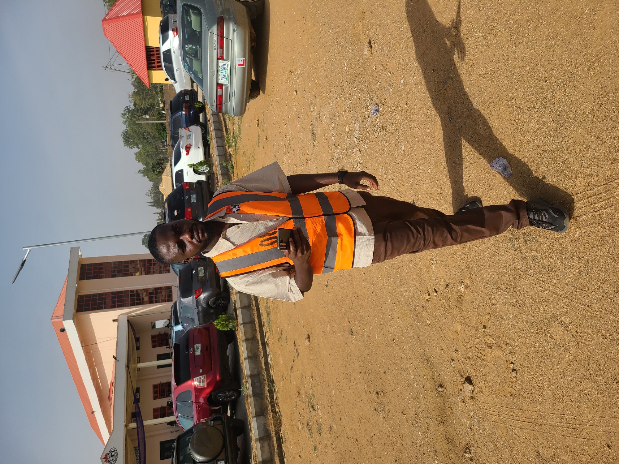 A man wearing an orange safety vest, brown trousers, and dark shoes stands on a sandy surface. He is holding a cell phone in his hand and has a watch on his wrist. His shadow is visible to his right. Behind him are parked cars and a building with a light-colored facade and a red roof. The license plate of the silver car reads PKN-118PN. The location is the city of Jos, Nigeria.