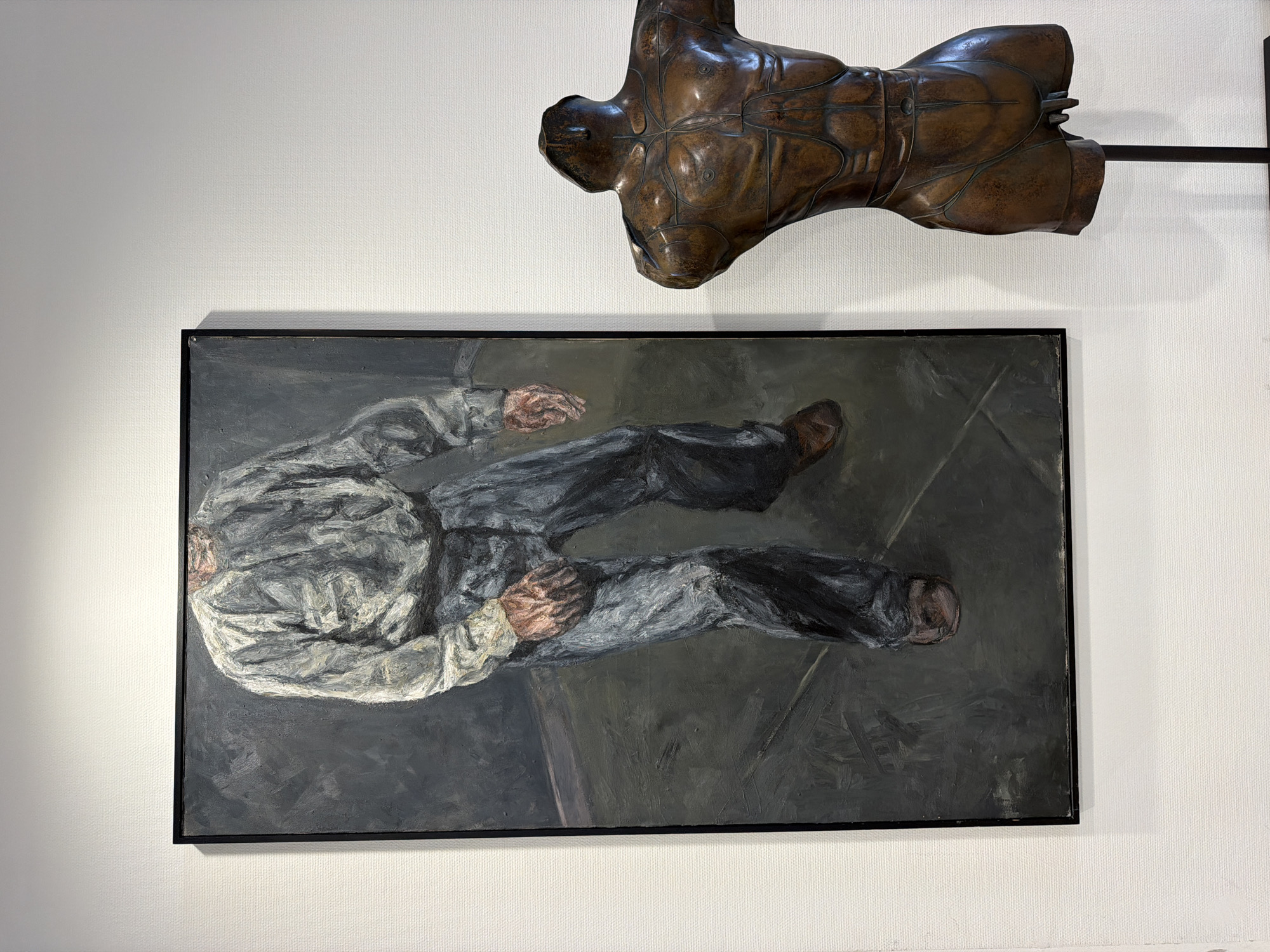The image displays two artworks mounted on a white, textured wall. On the left is a large, vertical painting framed in black, depicting a person from the upper torso down to the feet. The figure is wearing a loose, light-colored shirt and dark trousers, with brown shoes. The painting has a muted color palette, primarily grays and dark blues, with visible brushstrokes. To the right of the painting is a bronze sculpture of a male torso, mounted on a black stand. The sculpture is detailed, showing muscular definition and segmented lines across the chest and abdomen, with the head and limbs absent. The background wall is plain and white, providing a neutral setting for the art. The scene appears to be an exhibition or gallery space. The location is the Museum Bochum, in Bochum, Germany.