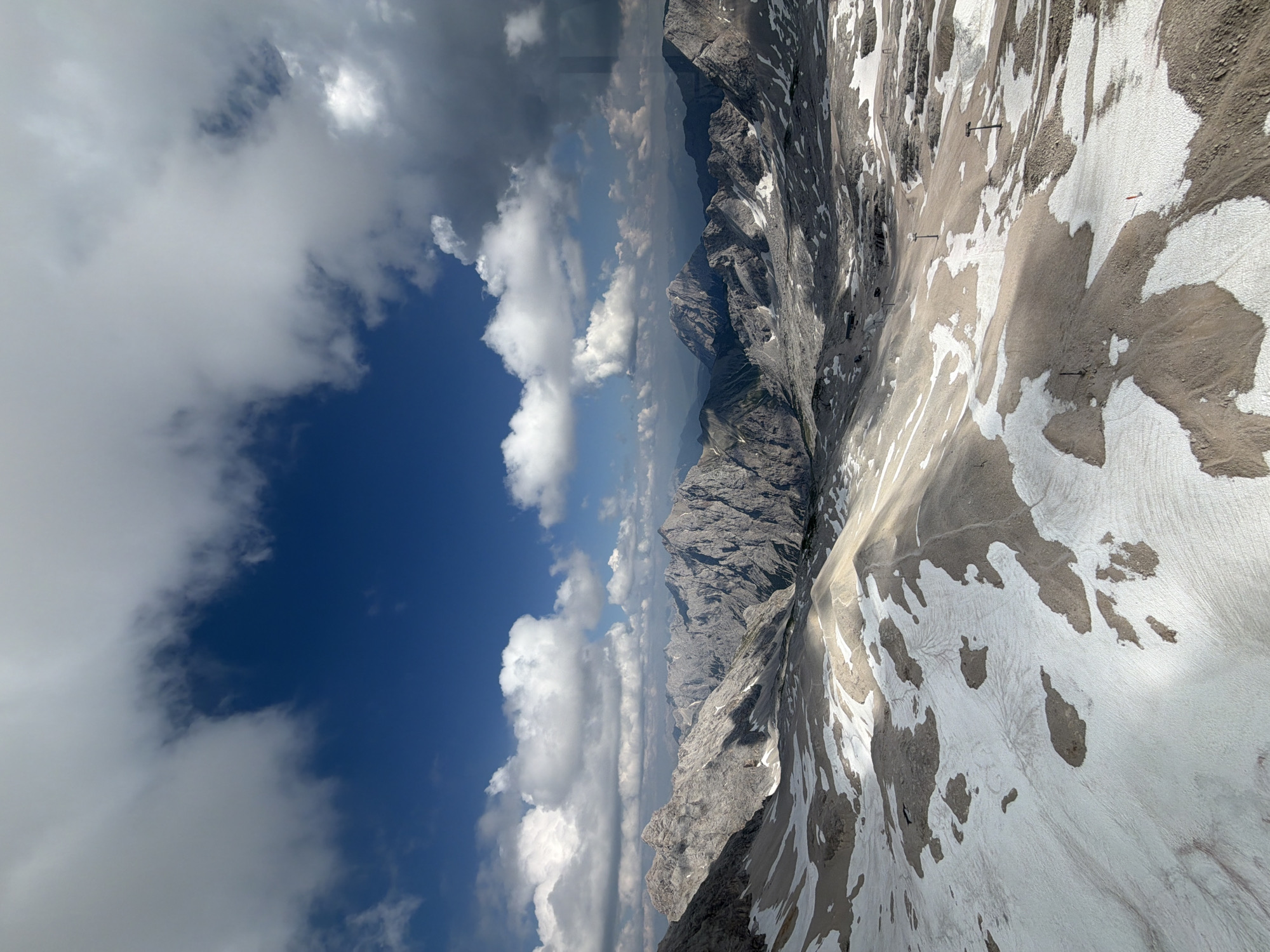 This image depicts a panoramic view of a mountainous alpine landscape with patches of snow and rock. The sky is a vibrant blue, punctuated by scattered white and gray clouds. The foreground is dominated by a snow-covered slope with areas of exposed rock and earth. In the middle ground and background, a series of rugged, rocky mountain peaks stretch into the distance, their surfaces textured by shadows and sunlight. A few chairlifts and poles are visible on the slopes, suggesting a ski resort or a popular hiking area. The overall scene conveys a sense of vastness and natural beauty. This is likely the Zugspitze, the highest peak in Germany, located in the Wetterstein range near Garmisch-Partenkirchen.