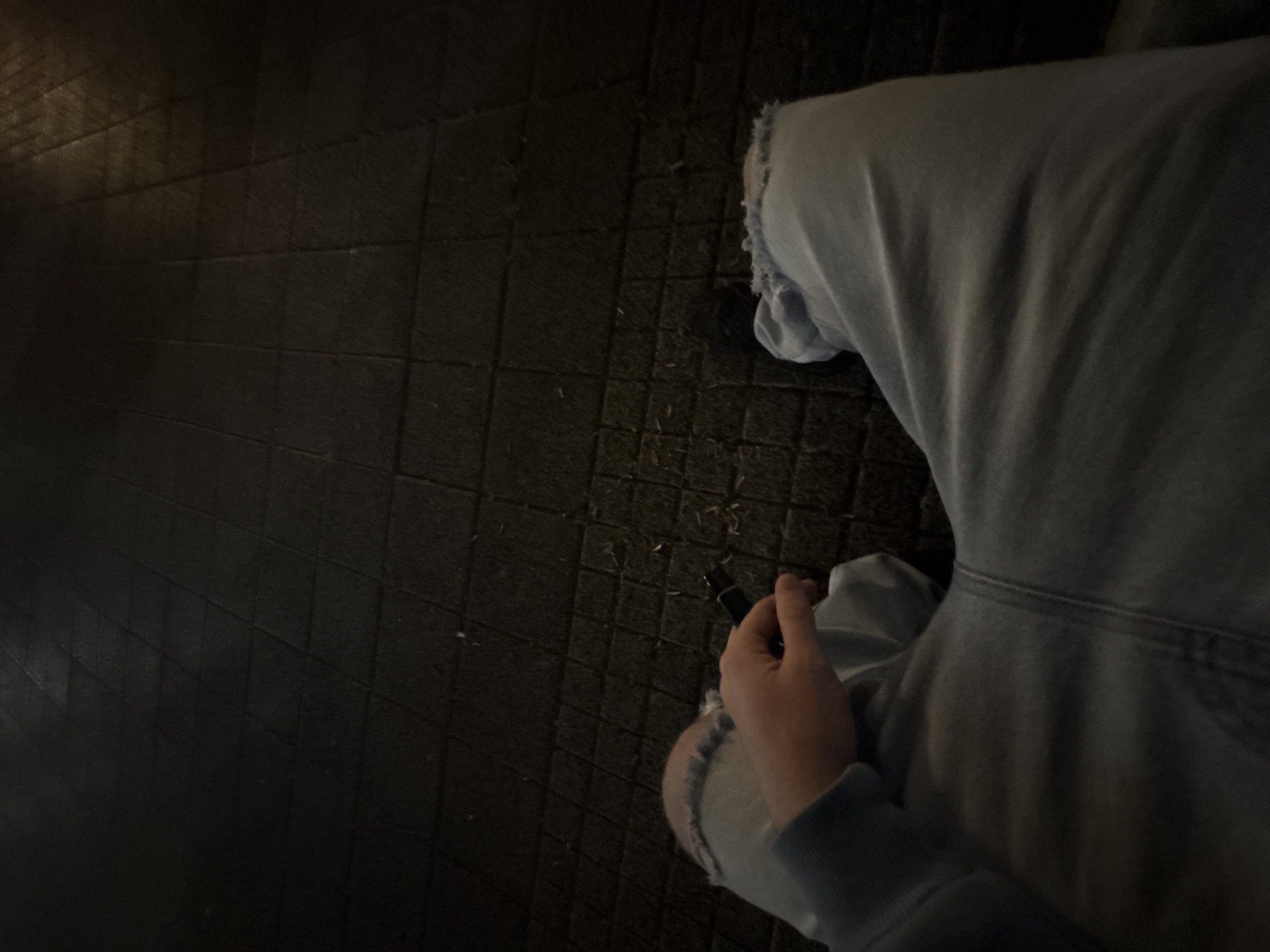 A low-angle, downward perspective captures a person's lower torso, legs, and right hand in a dimly lit setting, likely in Plovdiv, Bulgaria. The individual is wearing light-colored (possibly light blue or grey) jeans or trousers, which feature a distinct ripped detail on the right knee. Their right hand is visible, holding a slender, dark-colored object, which appears to be a pen, vape pen, or similar small device. The person's posture suggests they are either sitting or standing and looking downwards. The ground beneath them is a dark, textured surface, seemingly paved with square or rectangular tiles or blocks, and scattered with small, light-colored specks, possibly debris or lint. The overall lighting is low, indicative of either nighttime or an indoor environment with subdued illumination. Reflections of an unseen artificial light source are faintly visible on the left side of the frame. No discernible text is present in the image.