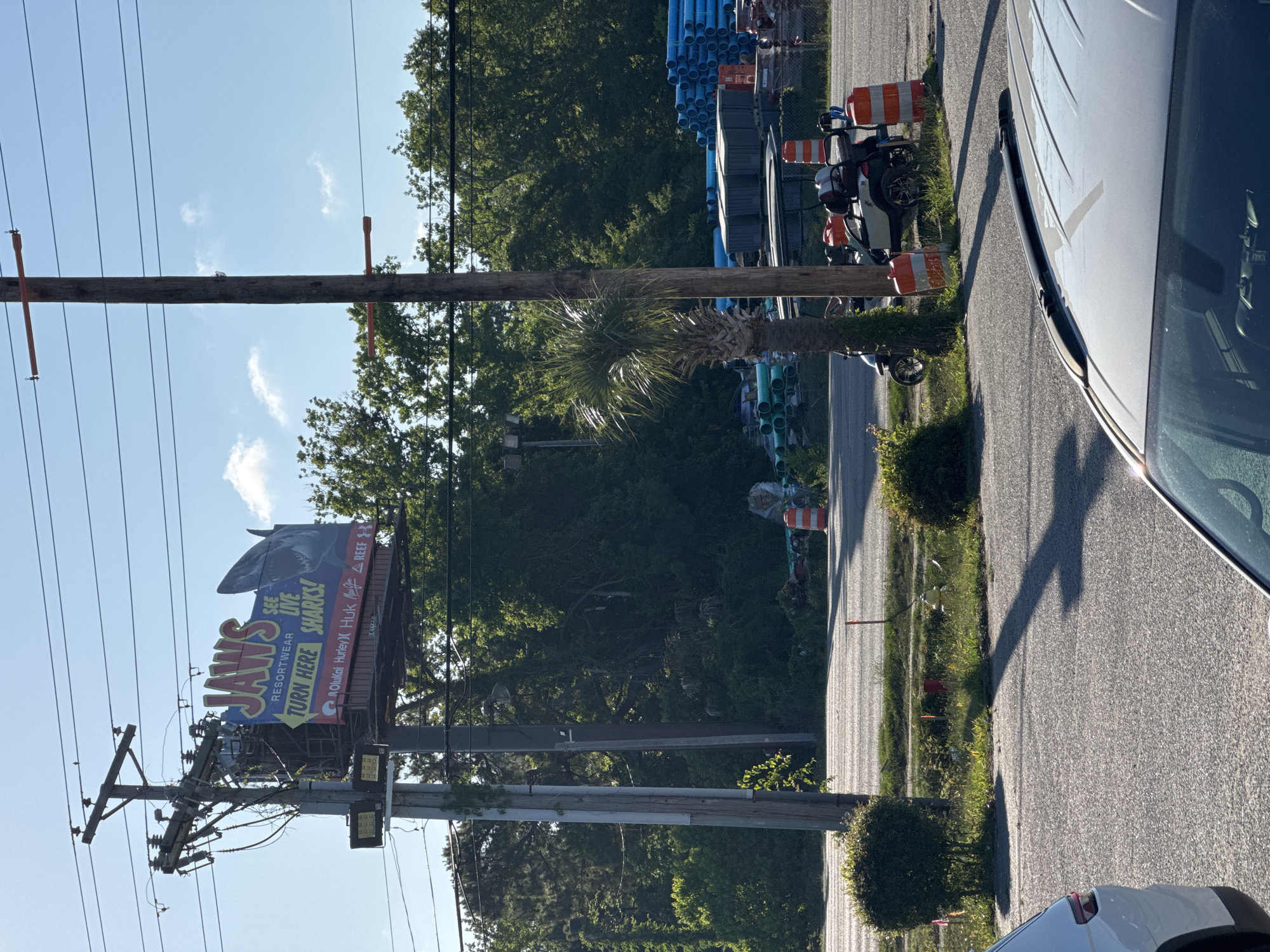 The image depicts an outdoor scene in Garden City Beach, United States of America. A large "JAWS Resortwear" billboard featuring a shark image is affixed to a utility pole, advertising live shark viewing and pointing towards a specific direction. The utility pole also carries electrical transformers and numerous power lines.
Adjacent to the road, construction materials, primarily stacks of blue PVC pipes, are visible behind a fence, suggesting an ongoing construction or utility project. Several orange and white striped traffic cones are also present, indicating road work or construction in the vicinity.
A palm tree is visible amidst the trees. A white vehicle is partially visible in the foreground and to the right side of the scene. The street appears paved and is likely part of the road network in Garden City Beach.