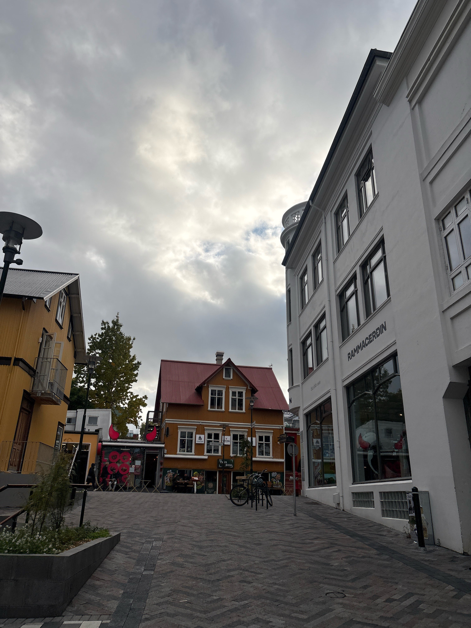 This is an eye-level, medium shot of a street scene in Reykjavik, Iceland, under a cloudy sky. The street is paved with dark and light grey interlocking stones arranged in a herringbone pattern. On the right is a multi-story white building with large windows. Above the ground floor windows, the words "RAMMACERDIN" are inscribed in black lettering. To the left of this building, a narrower street or alleyway leads to colorful buildings. The most prominent building on the left is a bright yellow, two-story structure with a dark grey metal roof. It features a balcony with a railing. Further back, an orange building with a red roof is visible, with windows and stairs on its exterior. In front of the orange building, a small cafe with outdoor seating is apparent. Several bicycles are parked on the street. The overall atmosphere suggests a commercial or residential area within a city.