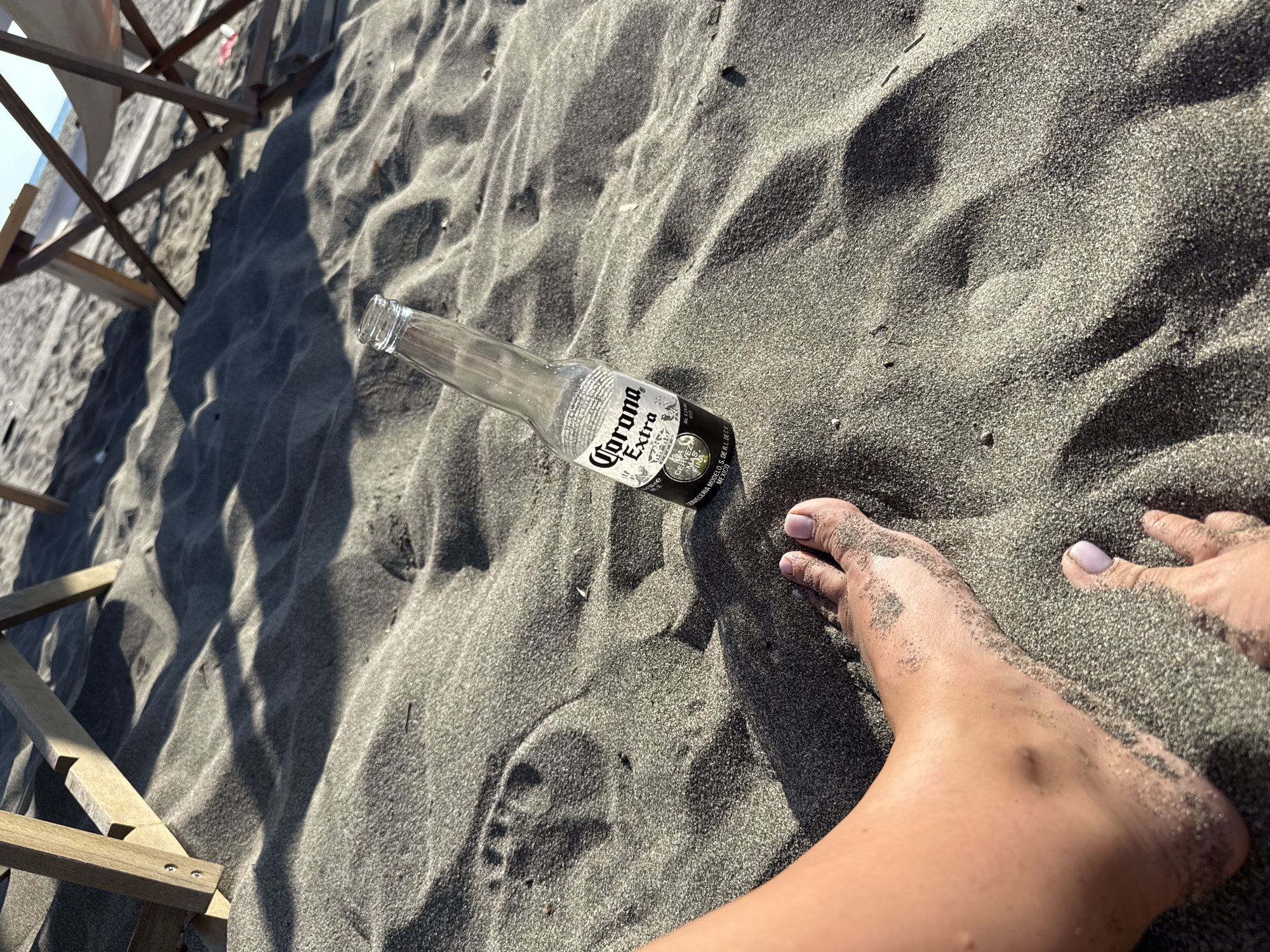 The photograph is taken on a dark sand beach in Gornji Štoj - Shtoji i Epërm, Montenegro. An empty, clear glass Corona Extra beer bottle lies on the sand. Adjacent to the bottle are a person's feet, covered in sand, with painted toenails. A partially visible wooden beach chair is located in the upper left corner of the image. The sand shows patterns from waves and wind.