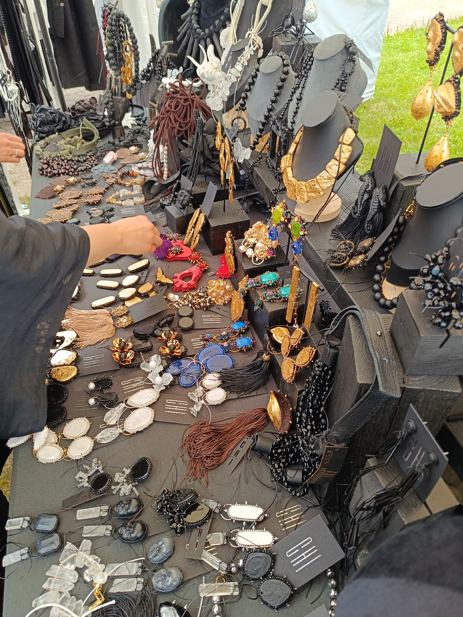 The image shows a close-up, eye-level view of a market stall displaying an assortment of handmade jewelry. The stall is draped with a dark fabric, likely a tablecloth, on which various necklaces, earrings, and pendants are laid out. The jewelry features a variety of materials including beads (black, white, and multi-colored), metal elements (gold and silver tones), natural stones, and what appear to be fabric tassels and decorative elements. Some pieces are displayed on black cardboard tags, and a few are mounted on small black busts. A person's hands, wearing a dark sleeve, are visible reaching out to examine some of the jewelry. In the background, there are more items on display, including clothing on hangers, and glimpses of greenery and possibly other market stalls. The overall impression is of a well-organized and aesthetically pleasing display of artisanal jewelry. This scene is characteristic of a market event in the historic town of Kuldīga, Latvia, known for its artisan crafts and vibrant market culture.