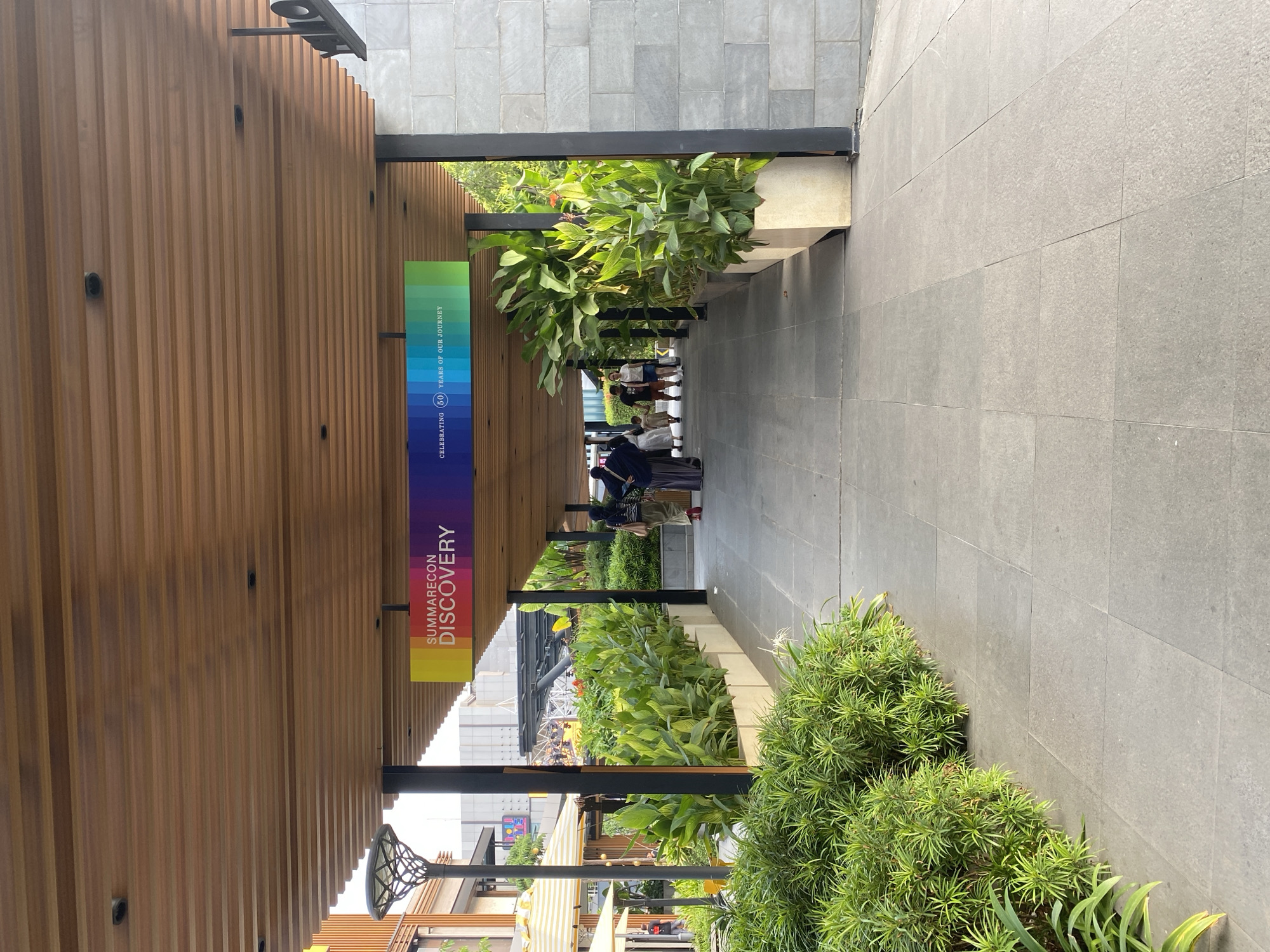 This image depicts a pathway at Summarecon Mall Kelapa Gading, located in North Jakarta, Indonesia. The pathway is covered by a wooden slatted ceiling, from which a banner displaying "SUMMARECON DISCOVERY" and "CELEBRATING 50 YEARS OF OUR JOURNEY" hangs. The banner features a colorful gradient. On either side of the pathway, there are planters filled with lush green plants. In the background, several people are walking along the pathway, some are dressed in traditional Islamic attire. The pathway is paved with large grey tiles. The architecture features modern design elements with a combination of wood and stone.