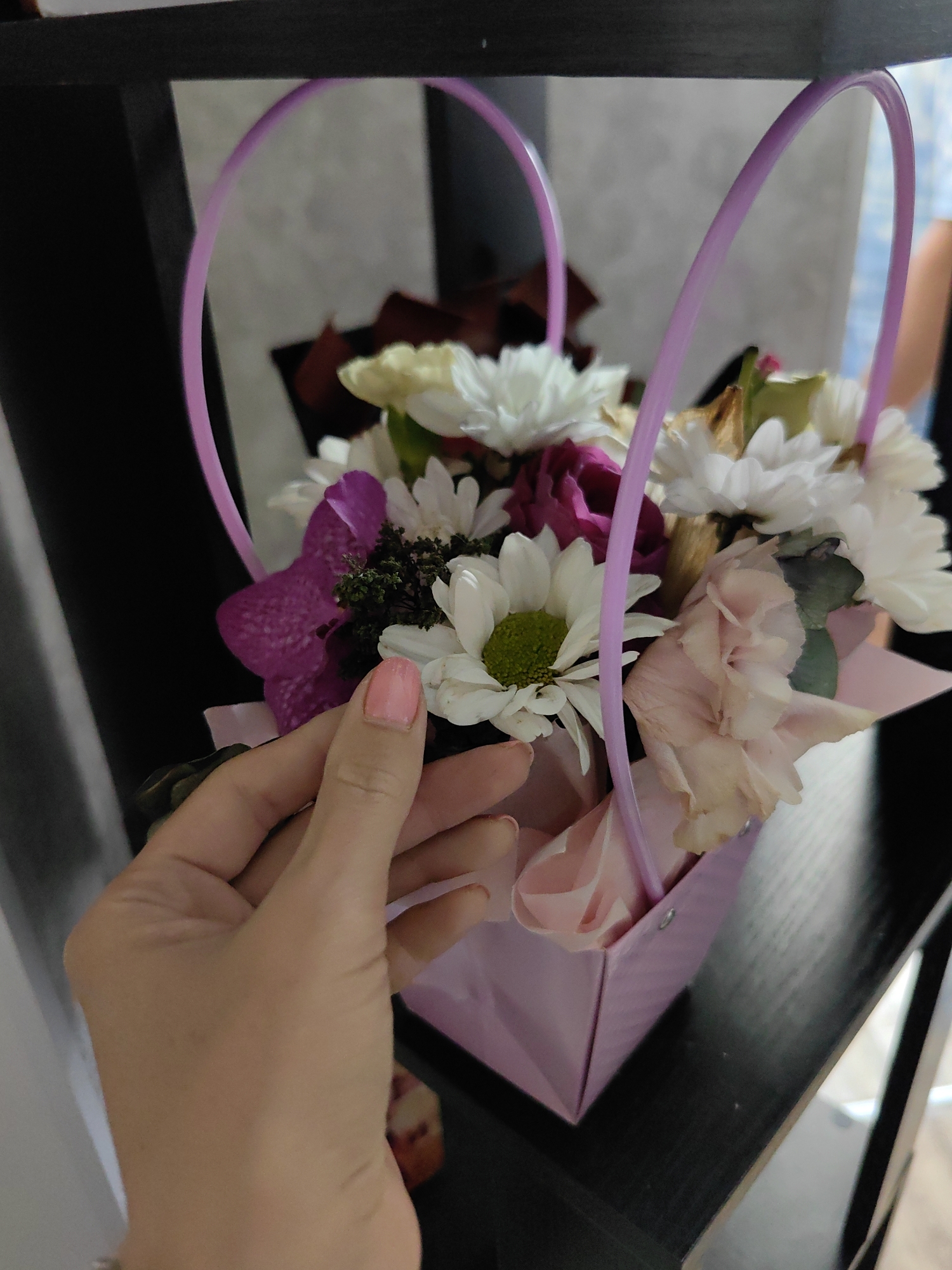 A close-up, top-down view shows a person's hand with pale pink nail polish gently touching a bouquet of flowers. The bouquet is arranged in a small, light pink paper gift box with two light purple loop handles extending upwards. The flowers are a mix of white chrysanthemums with green centers, deep pink, and pale pink roses, along with a prominent deep purple orchid. The background is blurred, with a dark shelf surface in the foreground and a lighter, textured wall behind. The image appears to be taken indoors. The location is not specified to be in the city of Myadzyel, Belarus.