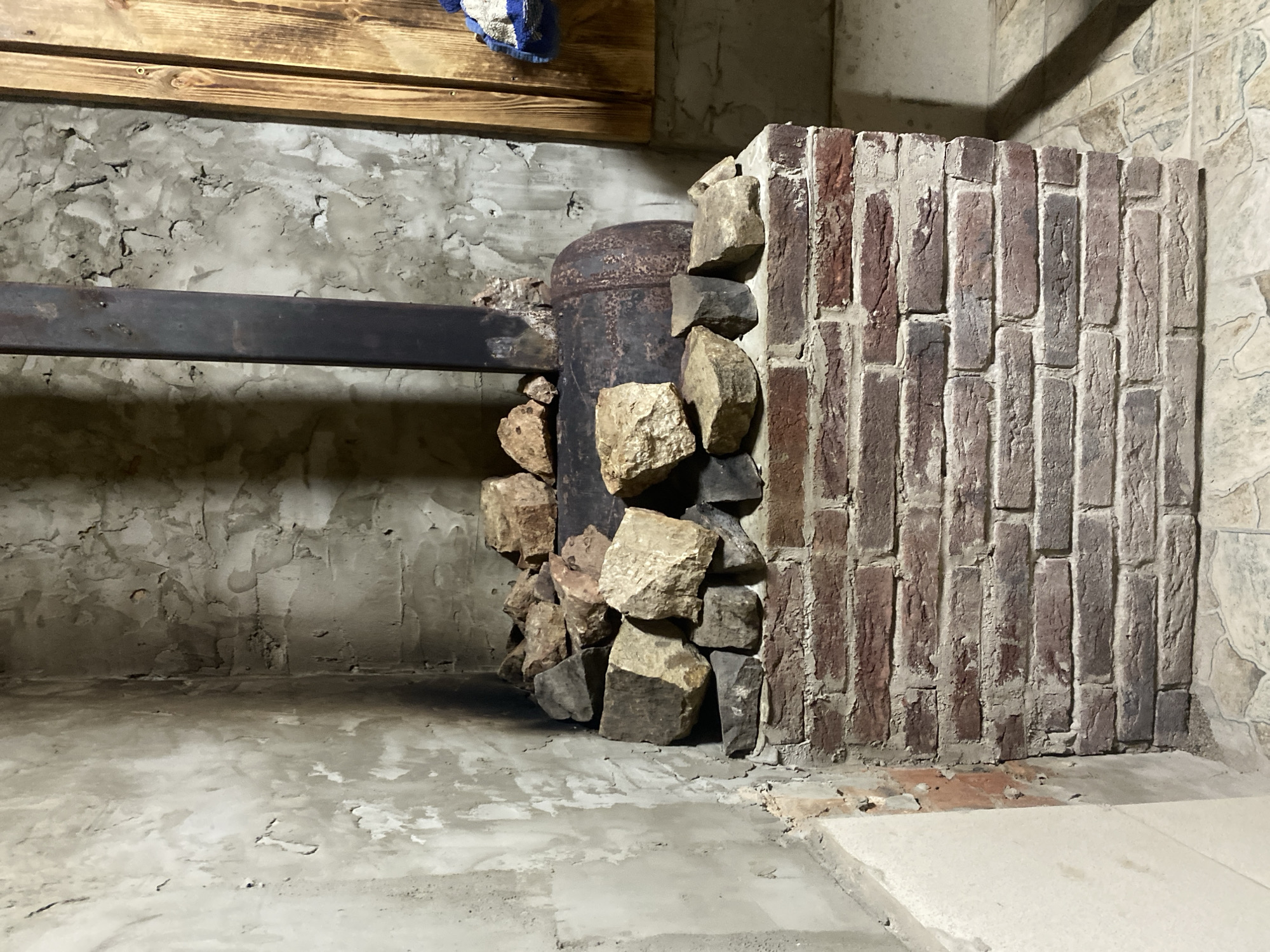 The image depicts a rustic sauna stove in what appears to be a basement or utility room. The stove is constructed with a round metal base, likely a repurposed tank, elevated on a brick pedestal. A dark metal chimney pipe extends vertically from the top of the stove. The stove and its base are adorned with a collection of rough, irregular stones, some of which appear to be heated stones commonly used in saunas. The walls surrounding the stove are roughly plastered and show signs of wear, with patches of discoloration and uneven texture. To the right of the stove, a section of wooden paneling is visible, and a blue and white towel hangs on it. The floor is tiled with irregularly shaped beige tiles. The overall impression is one of a functional, perhaps DIY, sauna setup in a simple, unadorned space. The location is the city of Kisliakovskaya, Russia. There are no people present in the image.
